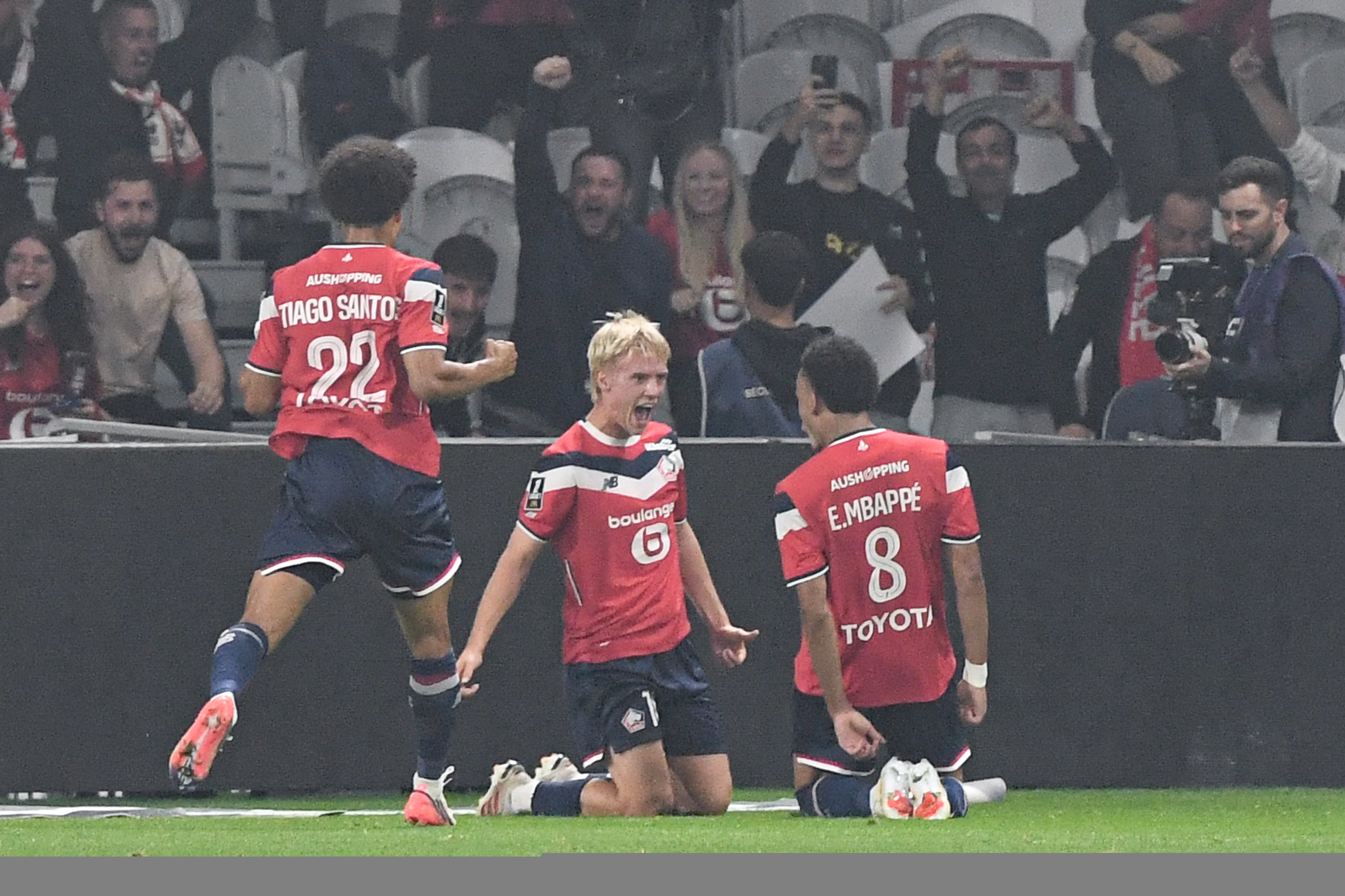 Fotbollsspelare i röd tröja firar tillsammans med lagkamrater på fotbollsplanen med publik i bakgrunden.