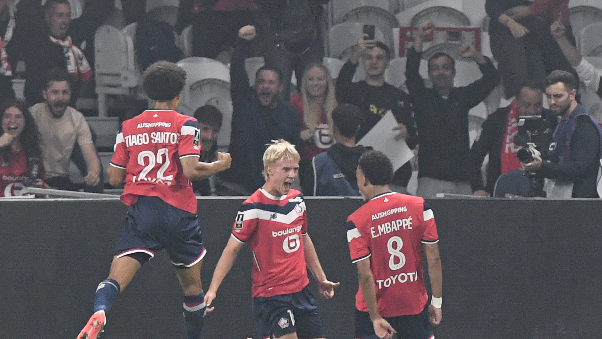 Fotbollsspelare i röd tröja firar tillsammans med lagkamrater på fotbollsplanen med publik i bakgrunden.