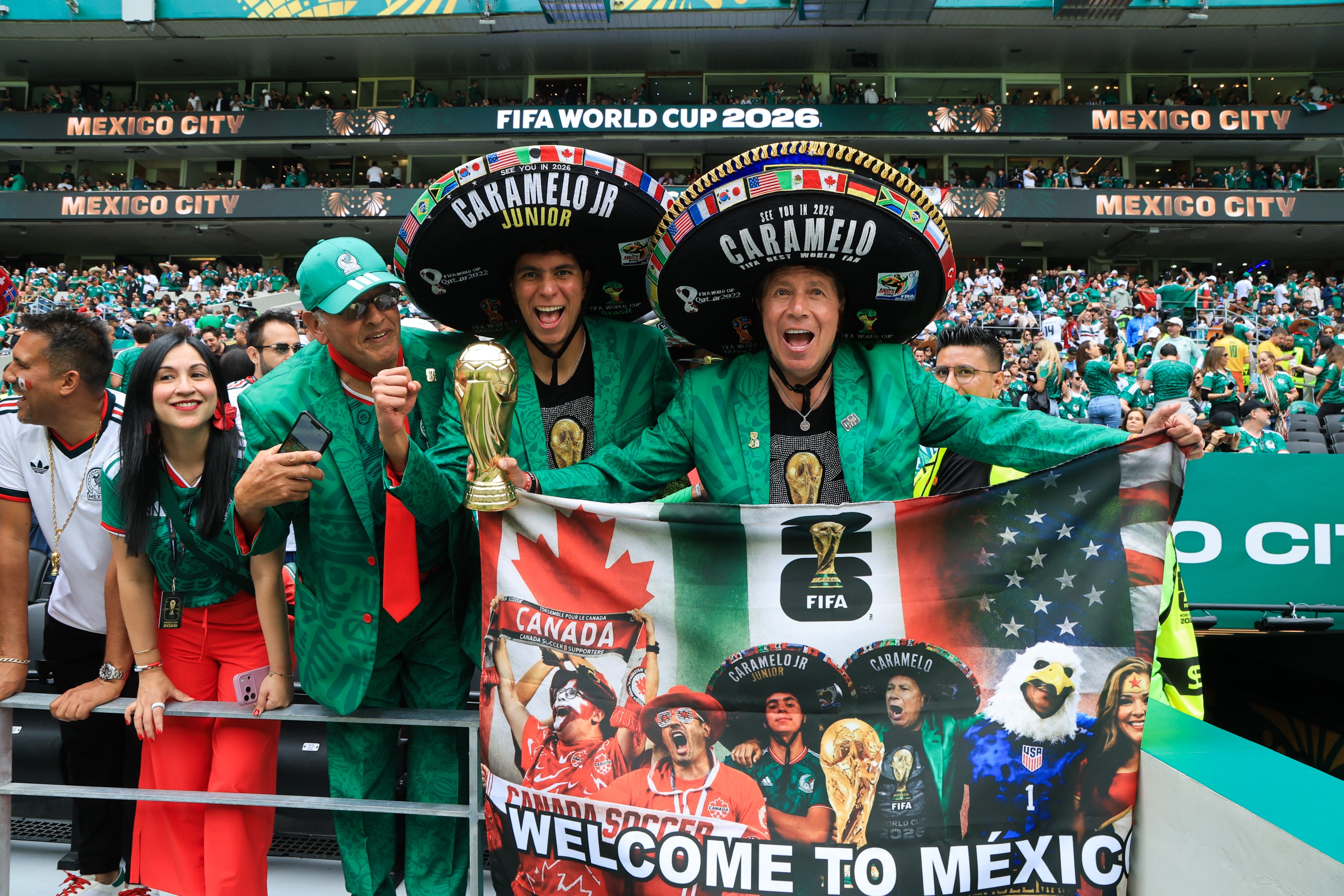 Mexikanska fotbollsfans i gröna och röda färger firar på läktaren under FIFA World Cup 2026 i Mexico City. De bär stora traditionella mexikanska hattar och håller upp Mexikos flagga och ett banderoll med texten 'Welcome to Mexico'. I bakgrunden syns fullt stadion med publik.