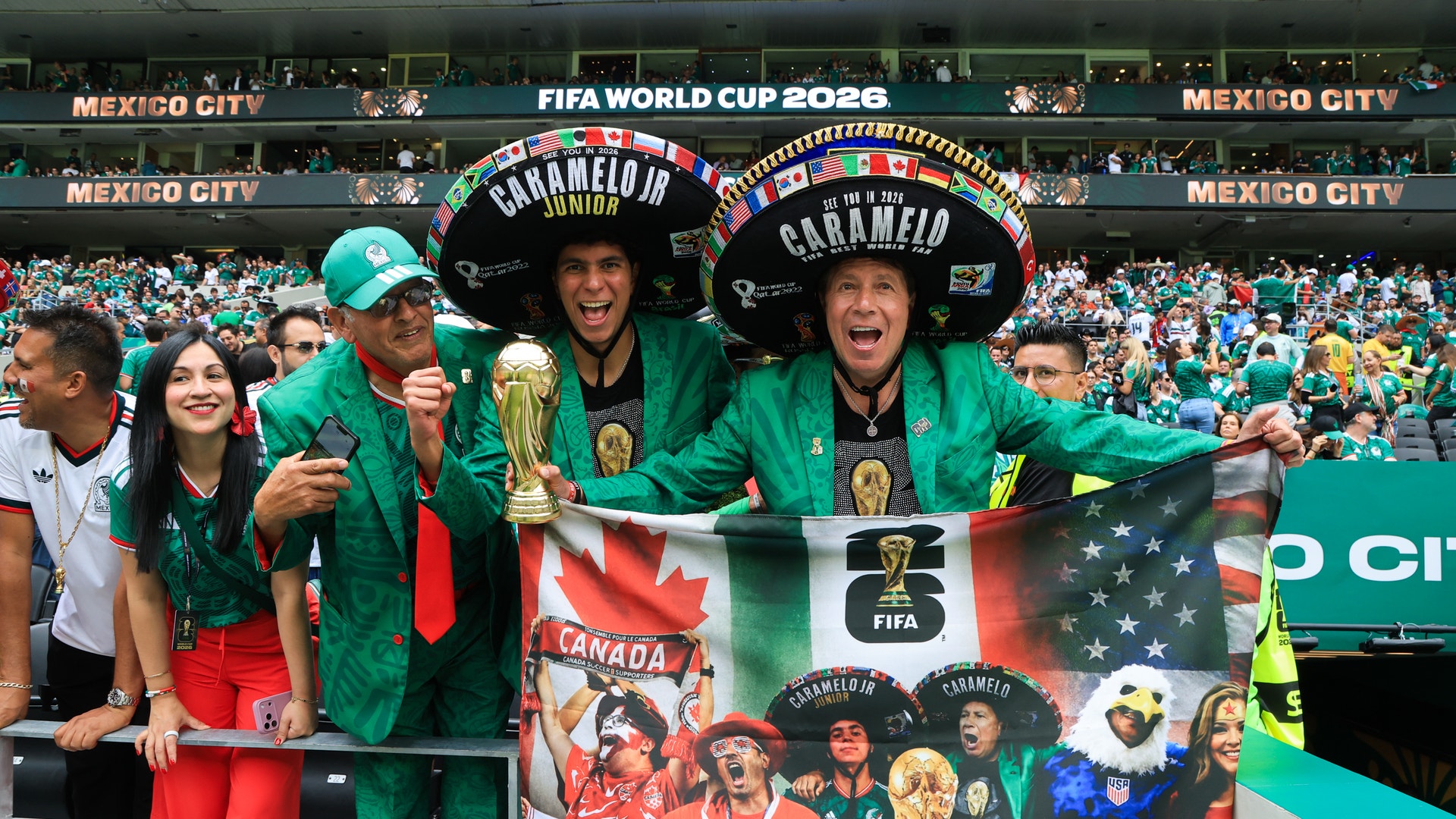 Mexikanska fotbollsfans i gröna och röda färger firar på läktaren under FIFA World Cup 2026 i Mexico City. De bär stora traditionella mexikanska hattar och håller upp Mexikos flagga och ett banderoll med texten 'Welcome to Mexico'. I bakgrunden syns fullt stadion med publik.