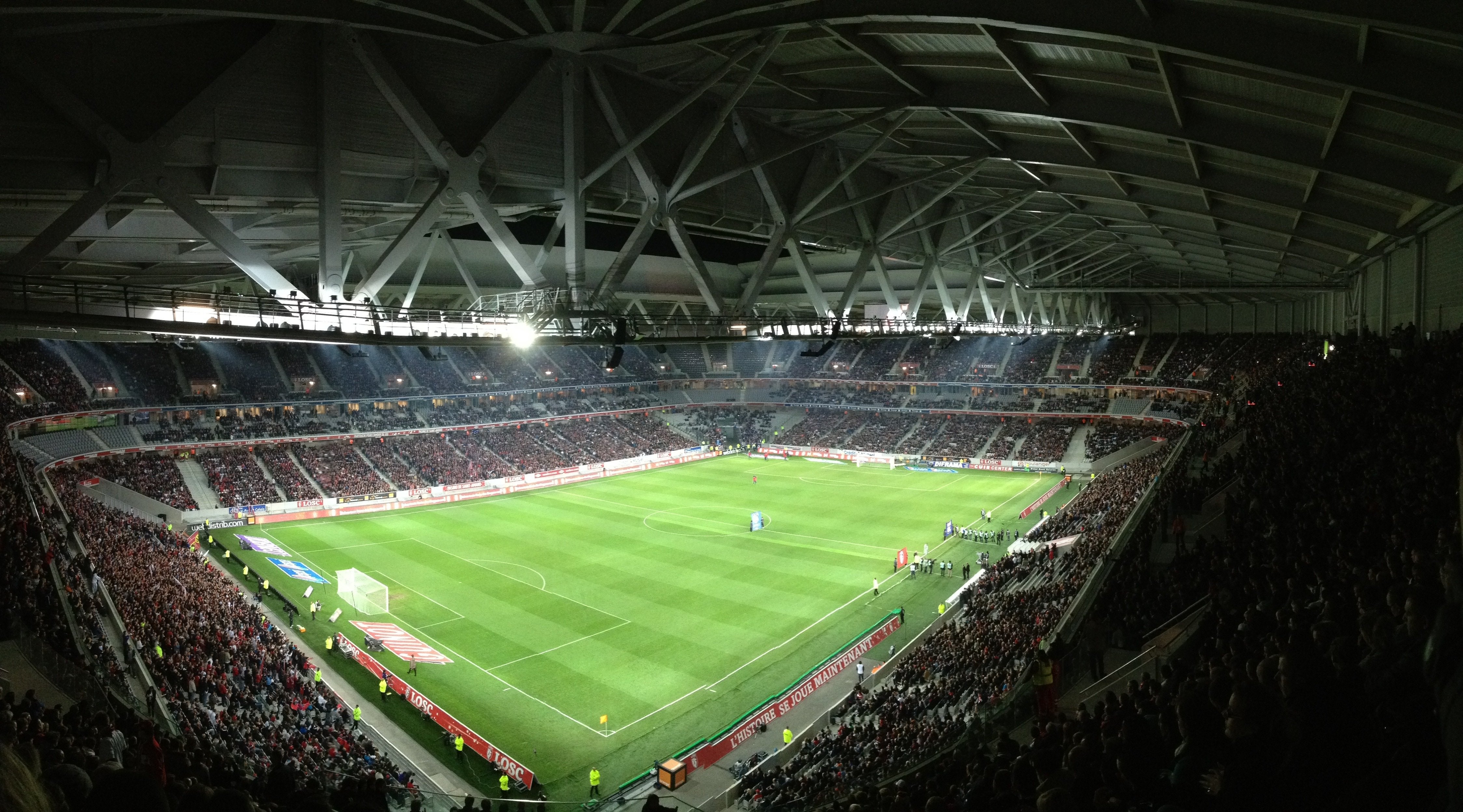 En stor fotbollsstadion fylld med åskådare under en fotbollsmatch. Gröna fotbollsplanen är väl upplyst och omgiven av moderna stadionkonstruktioner med metallkonstruktioner och belysning.