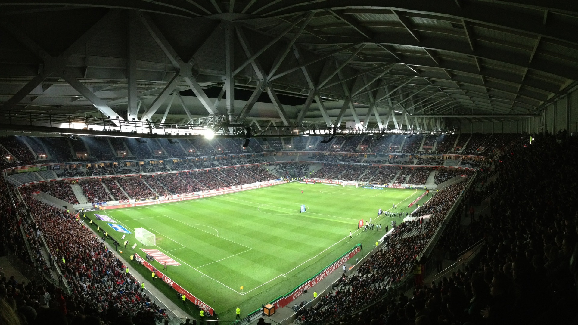 En stor fotbollsstadion fylld med åskådare under en fotbollsmatch. Gröna fotbollsplanen är väl upplyst och omgiven av moderna stadionkonstruktioner med metallkonstruktioner och belysning.