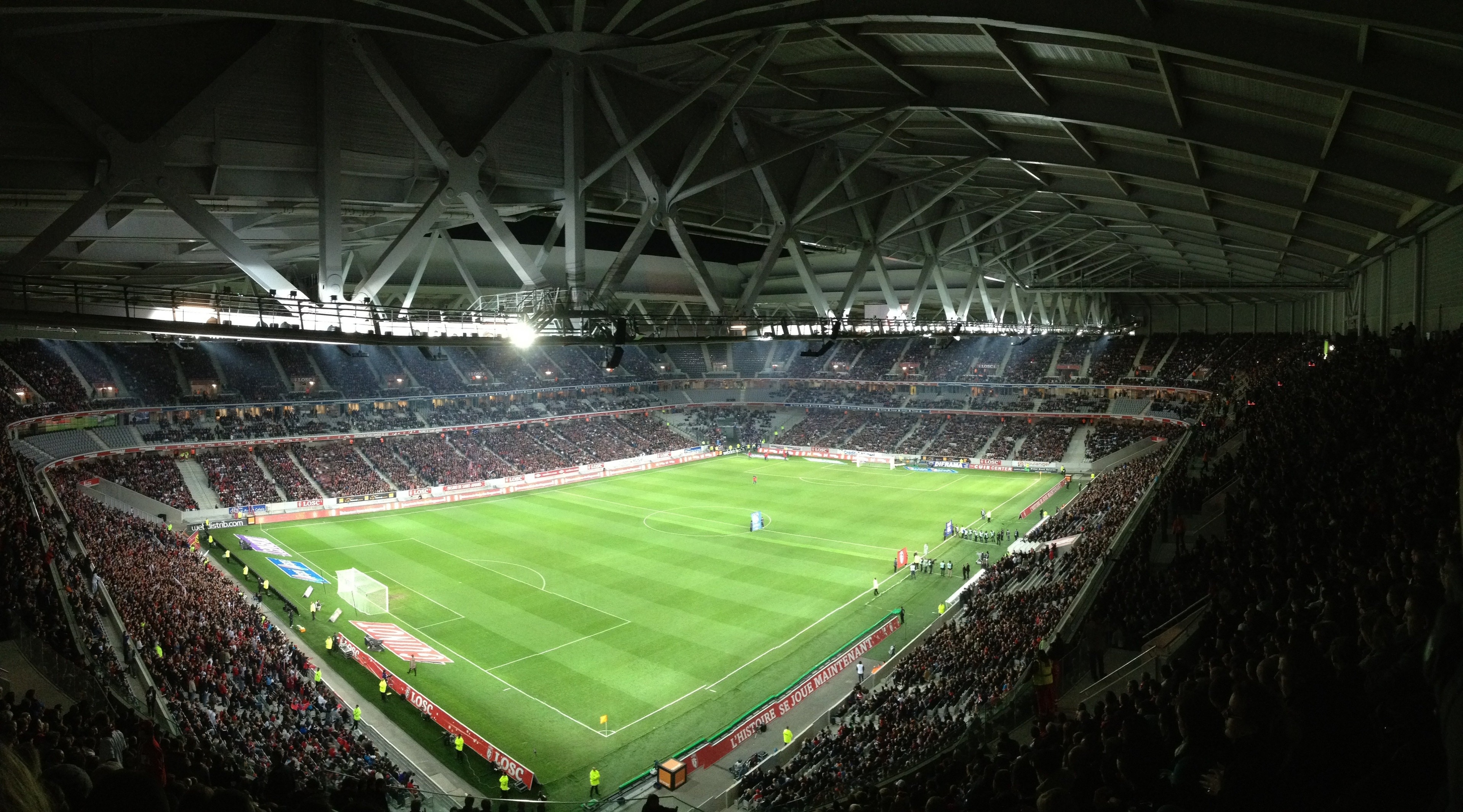 En stor fotbollsstadion fylld med åskådare under en fotbollsmatch. Gröna fotbollsplanen är väl upplyst och omgiven av moderna stadionkonstruktioner med metallkonstruktioner och belysning.