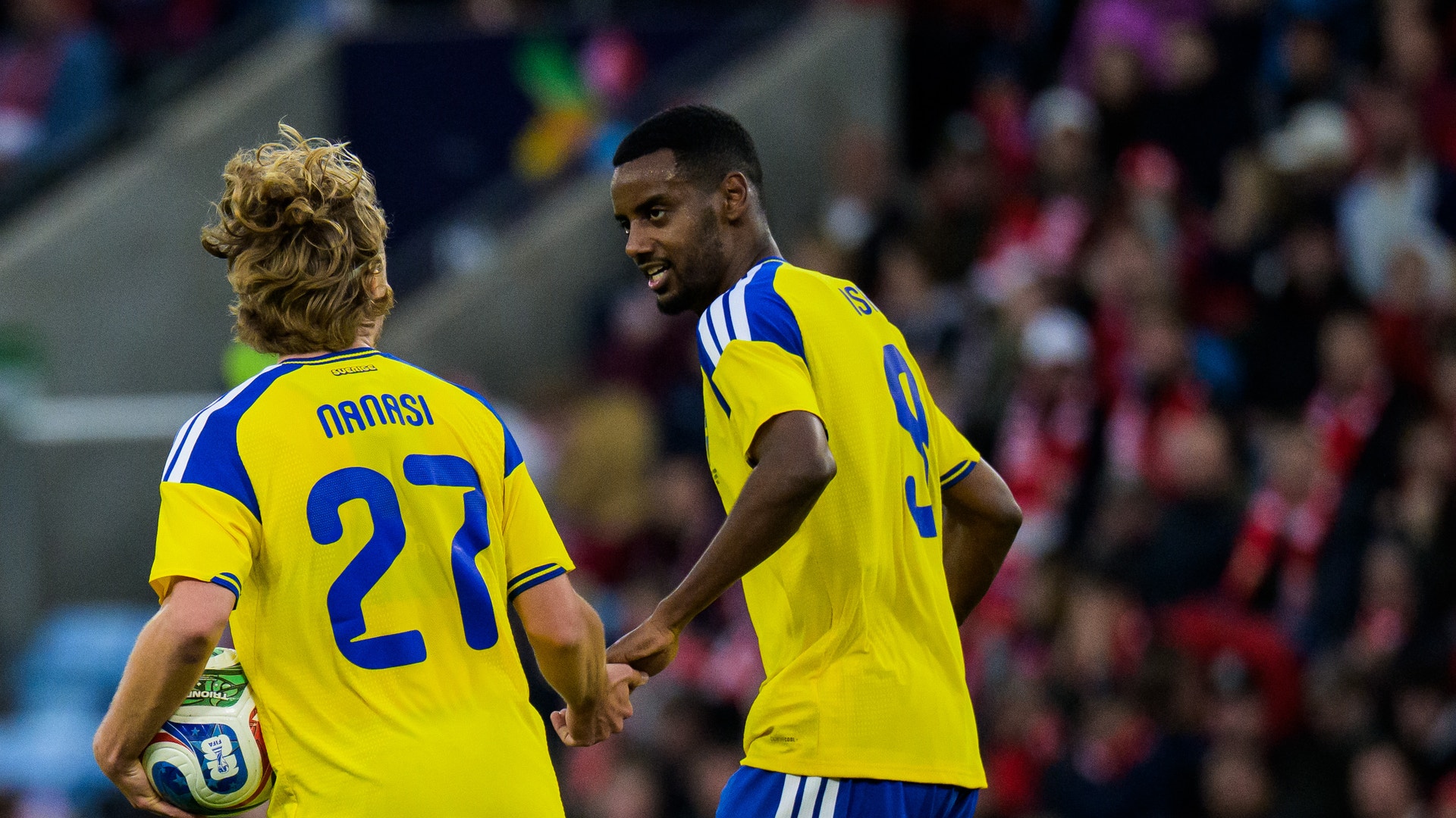 Två fotbollsspelare i svenska landslagströjor kommunicerar under en match på en stadion med publik i bakgrunden.