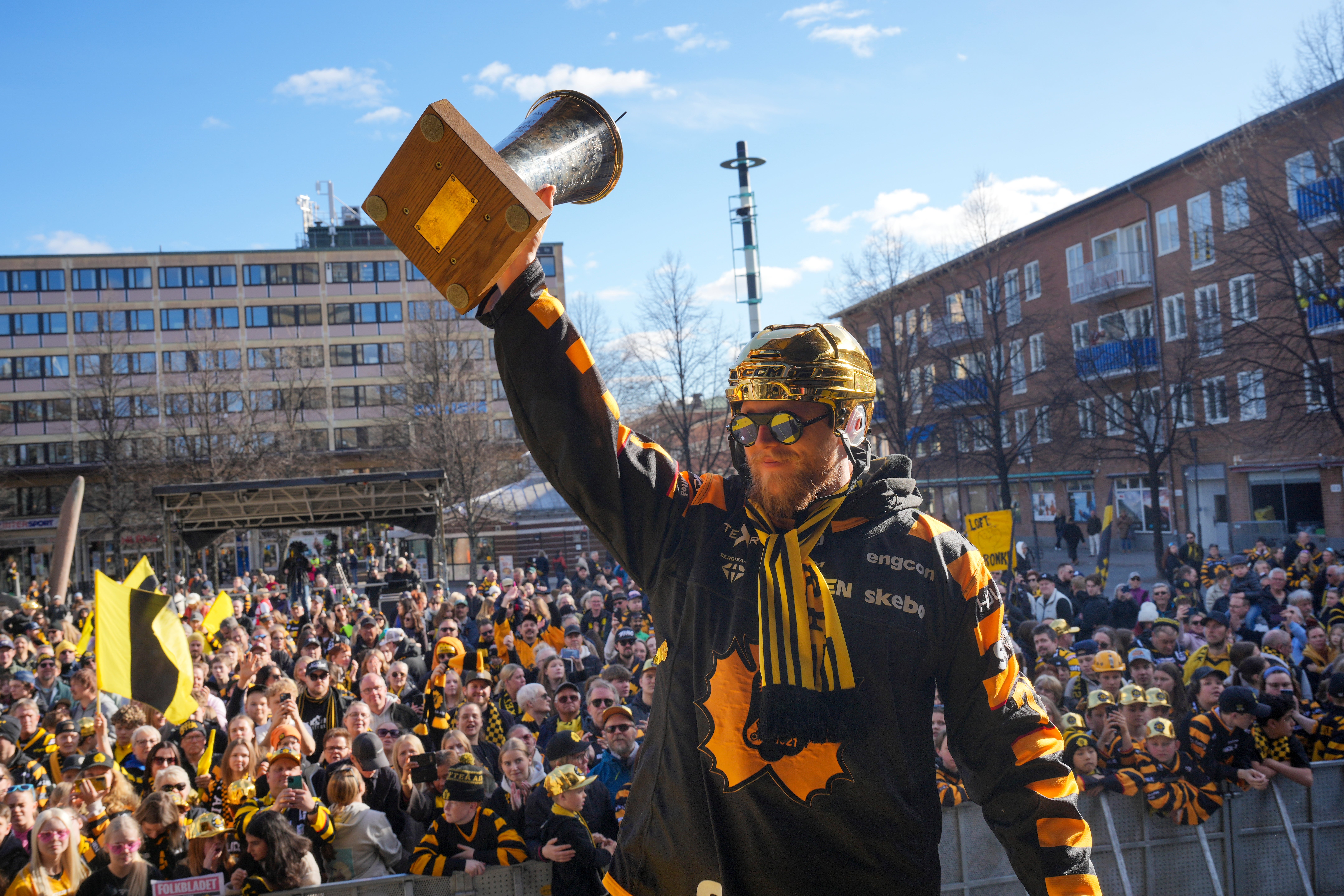 En person i gul och svart kläder håller upp en pokal framför en stor jubilande folkmängd på en stadsterrass i Skellefteå.