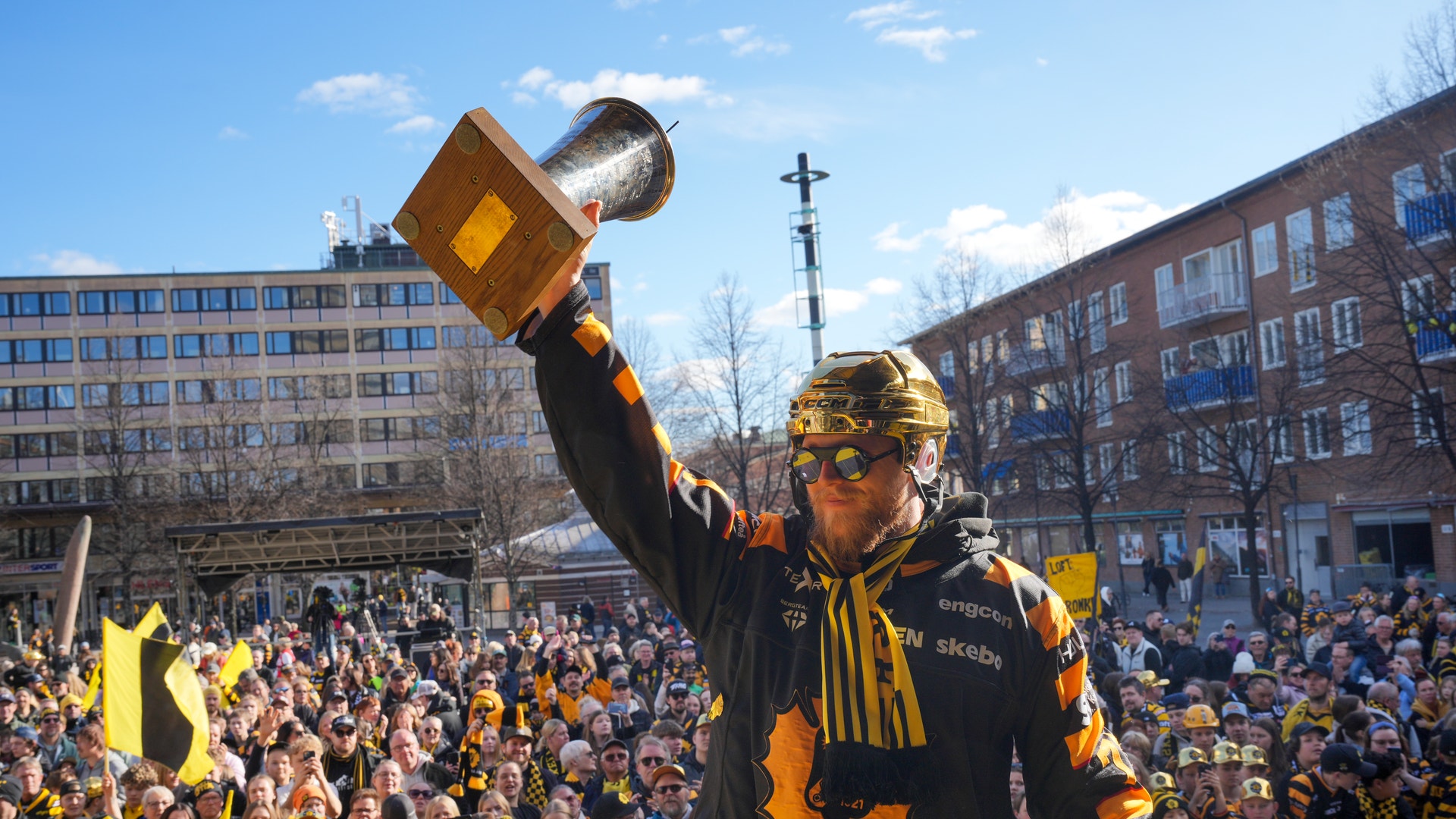 En person i gul och svart kläder håller upp en pokal framför en stor jubilande folkmängd på en stadsterrass i Skellefteå.