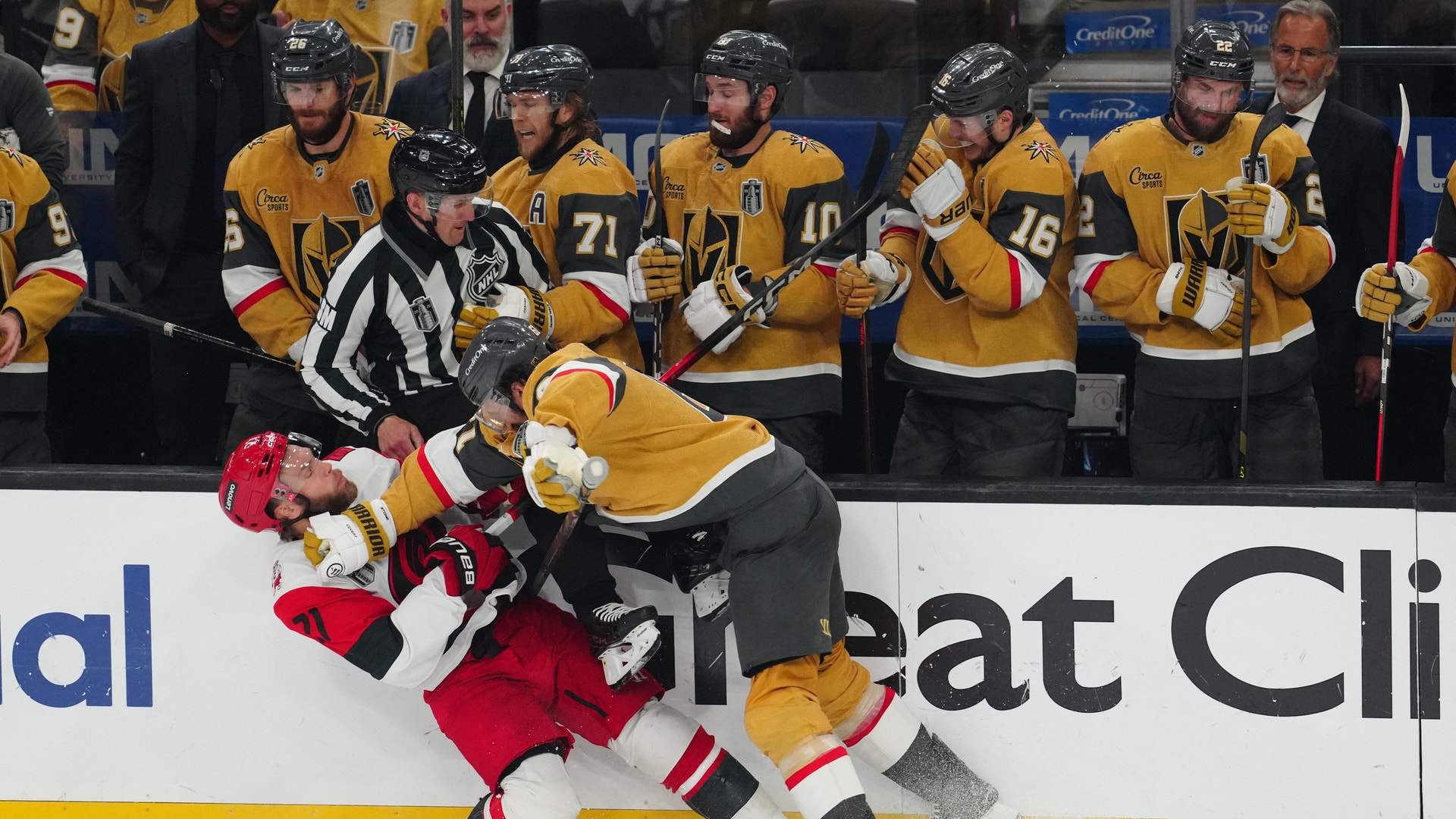 Ishockeymatch där en spelare i gul tröja tacklar en motspelare i röd tröja vid sargen. En domare övervakar spelet.