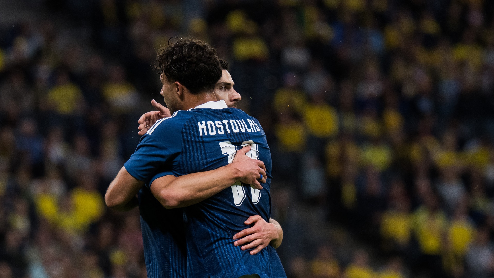 Två fotbollsspelare i blå tröjor omfamnas under en match med publik i bakgrunden.