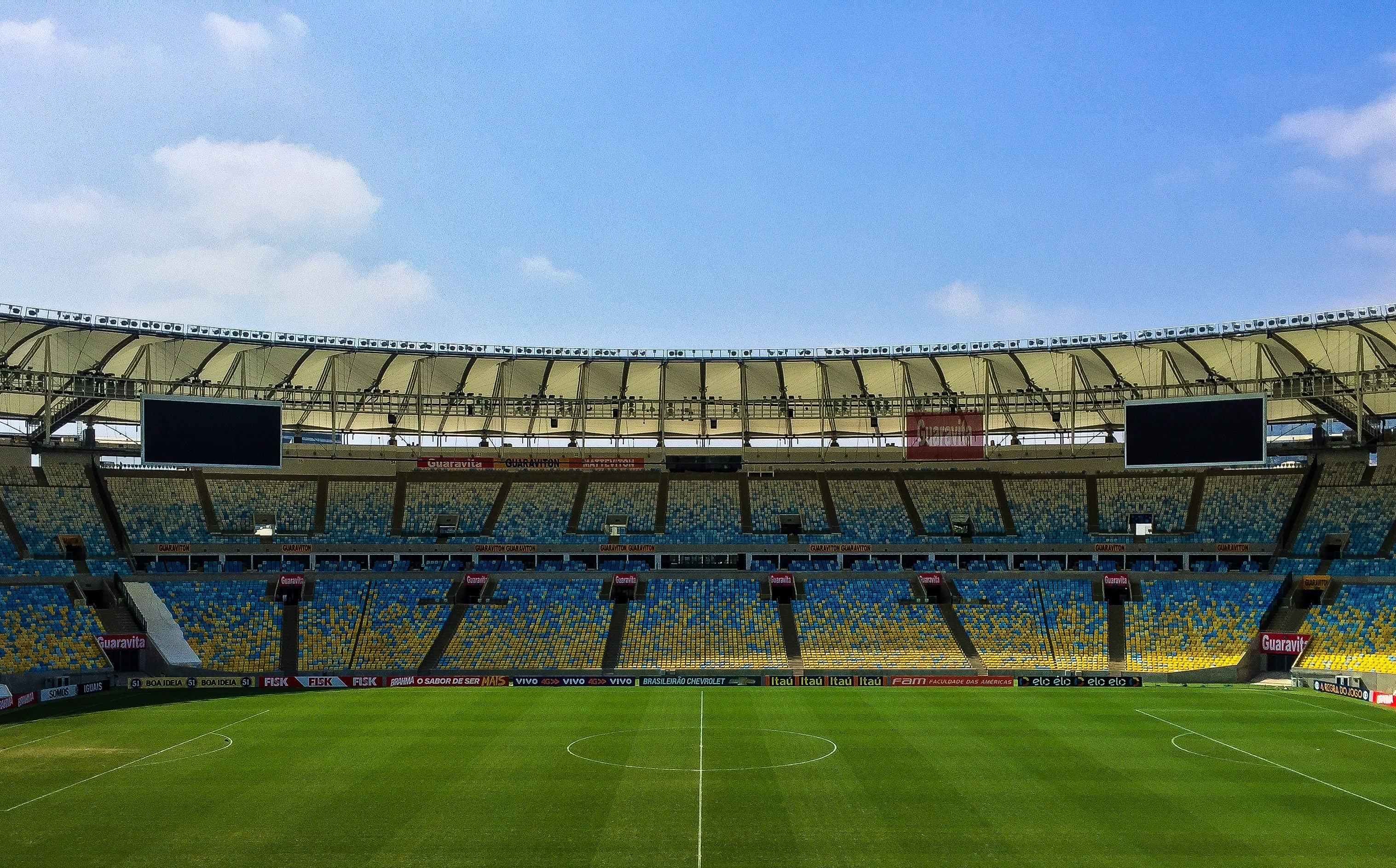 En stor fotbollsstadion med grönt gräs och vita mittlinjer. Läktarna är blåa och gula med ett modernt tak. Stadionen är tom och verkar vara under dagsljus.