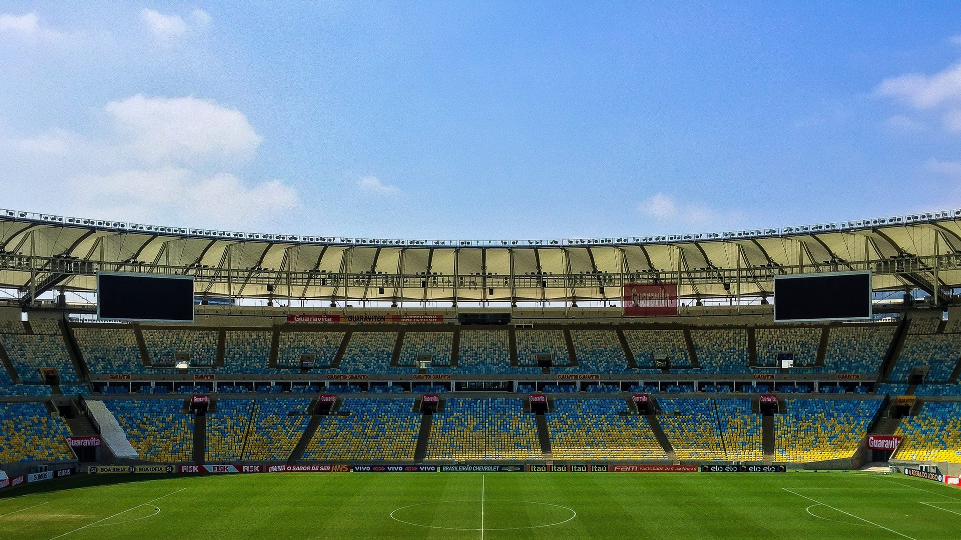 En stor fotbollsstadion med grönt gräs och vita mittlinjer. Läktarna är blåa och gula med ett modernt tak. Stadionen är tom och verkar vara under dagsljus.