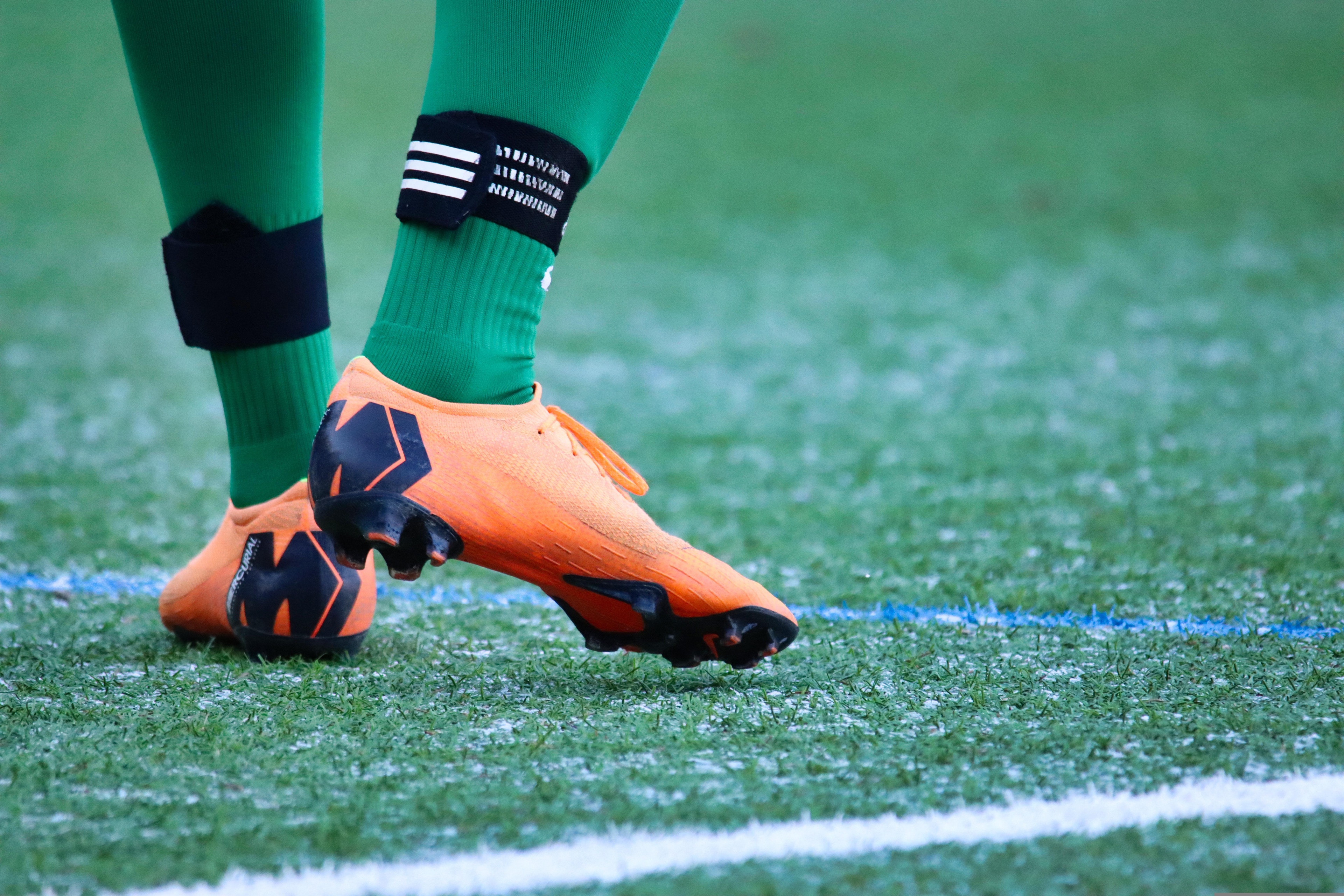 Närbild av en fotbollsspelares fötter i orange fotbollsskor och gröna strumpor på en fotbollsplan med vita linjer.