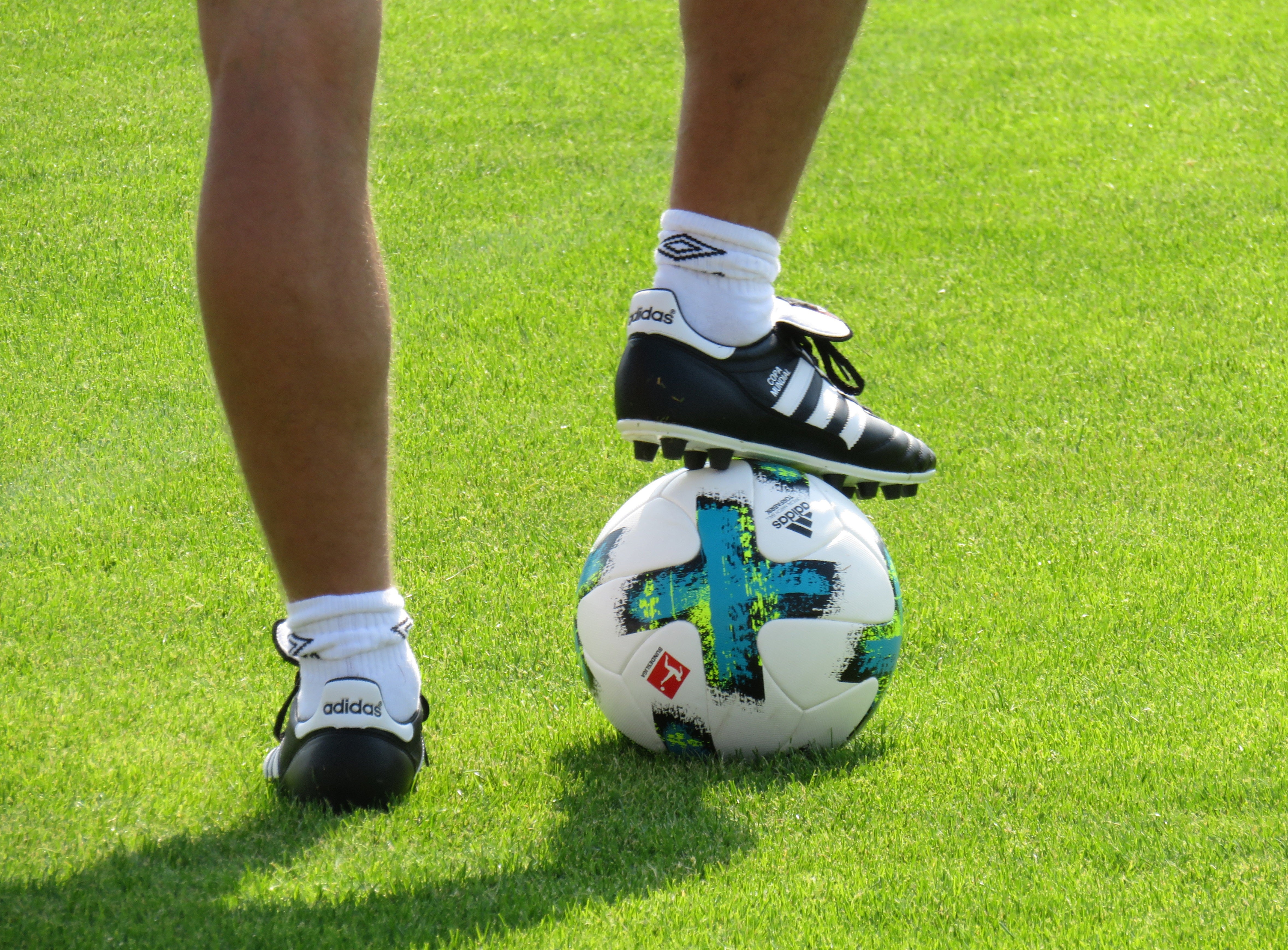 En fotbollsspelare i vita strumpor och svarta fotbollsskor står på en fotboll på en grön gräsplan.
