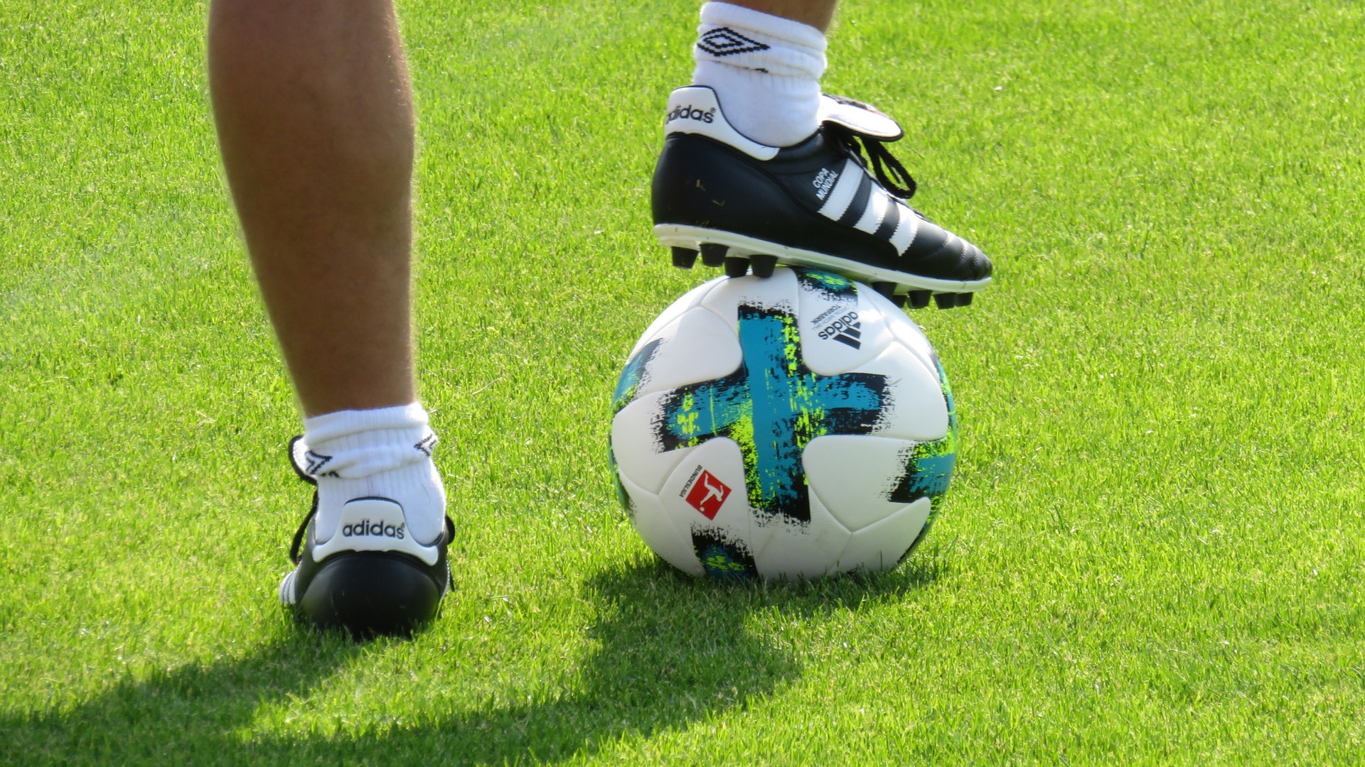 En fotbollsspelare i vita strumpor och svarta fotbollsskor står på en fotboll på en grön gräsplan.