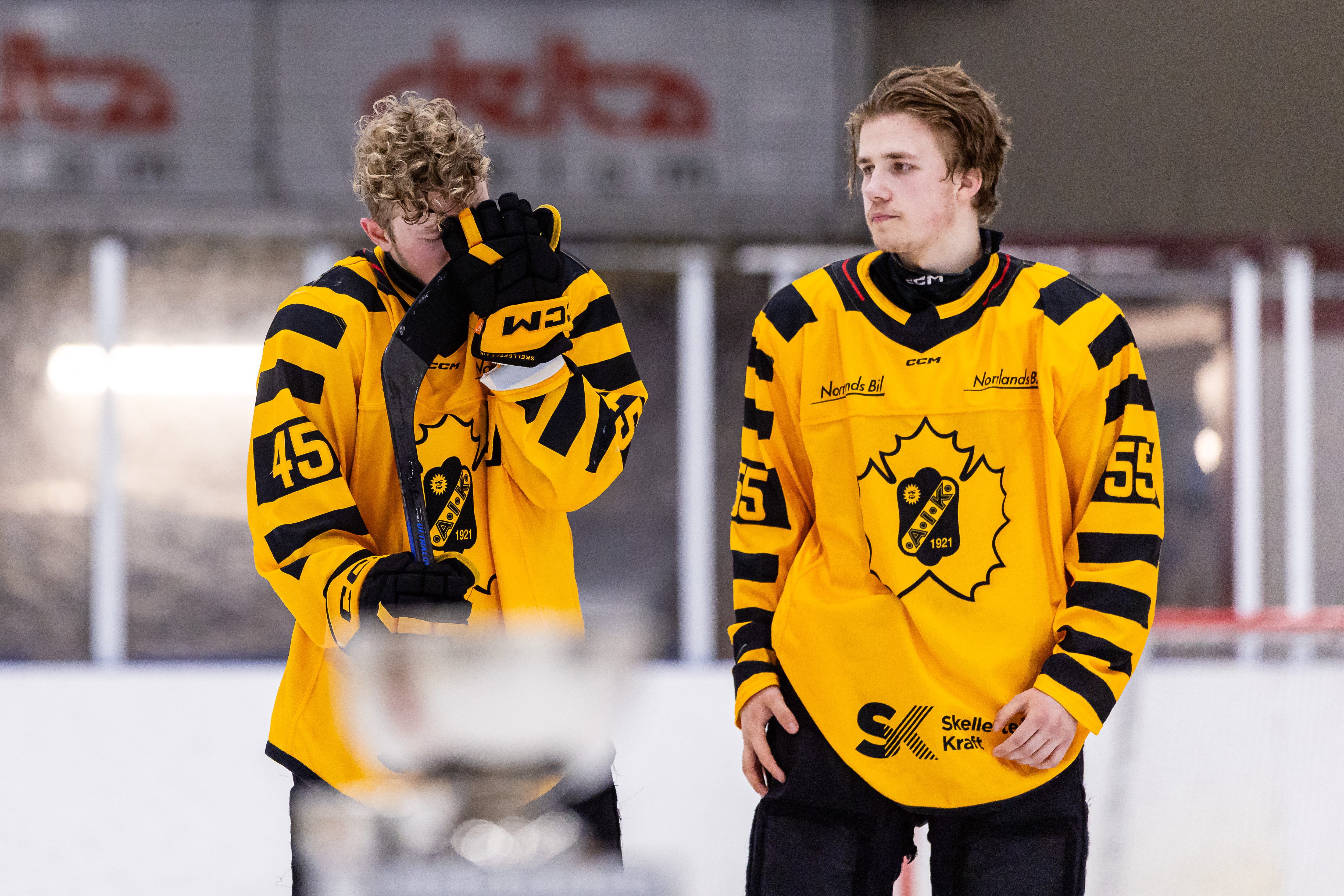 Två ishockeyspelare i gula och svarta tröjor står på isen under en match. Spelaren till vänster håller sin hockeyklubb och ser åt sidan, medan spelaren till höger står bredvid och tittar framåt.