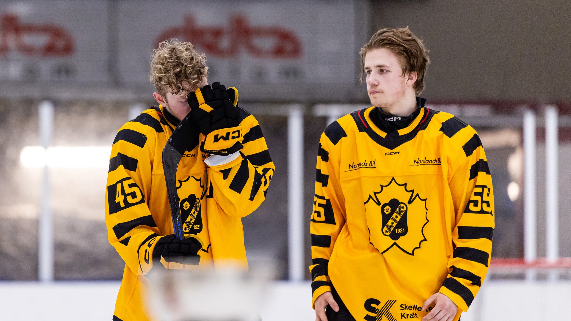 Två ishockeyspelare i gula och svarta tröjor står på isen under en match. Spelaren till vänster håller sin hockeyklubb och ser åt sidan, medan spelaren till höger står bredvid och tittar framåt.
