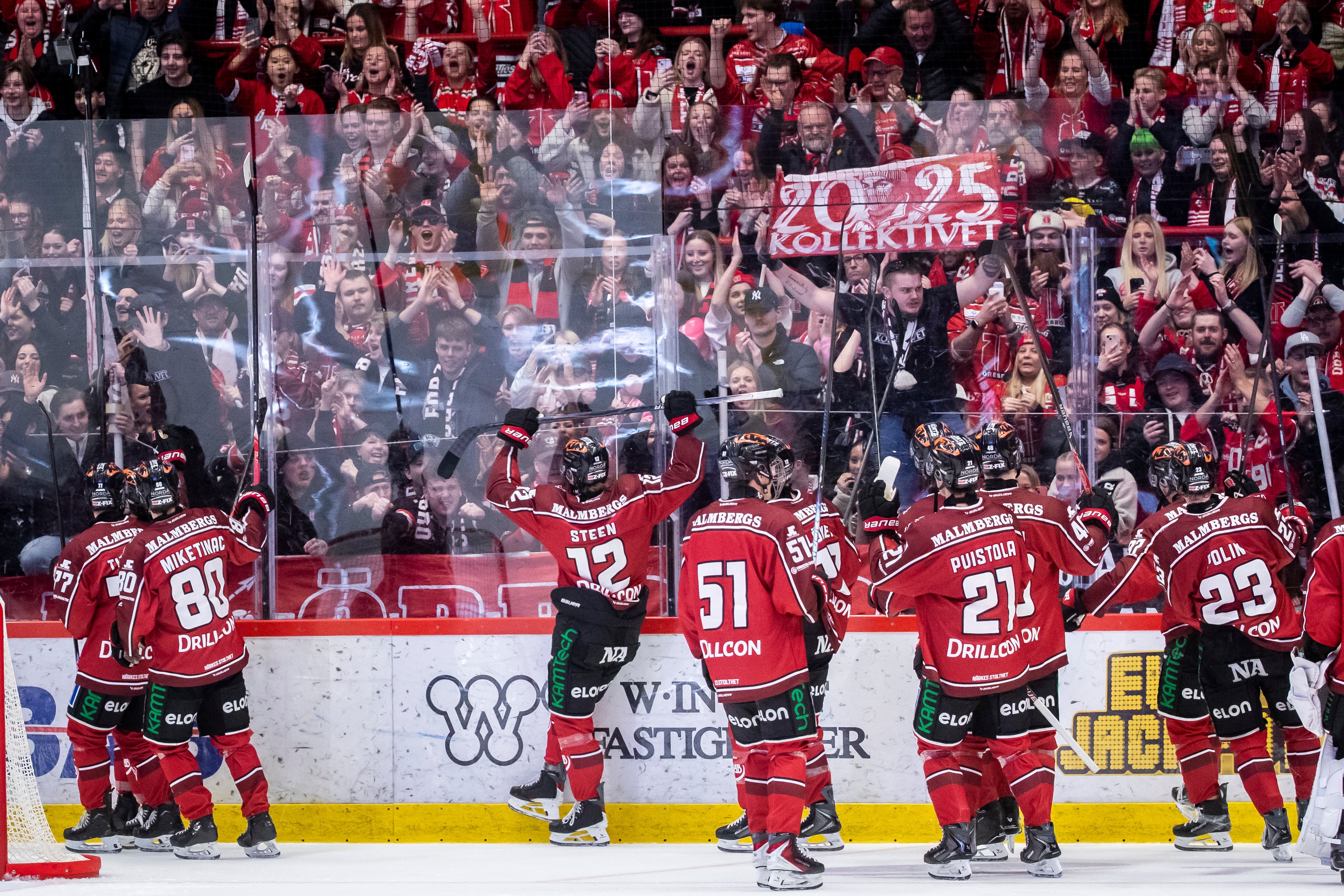 Ishockeyspelare i röda tröjor står längs sargen under en match med en full läktare av supportrar i bakgrunden.