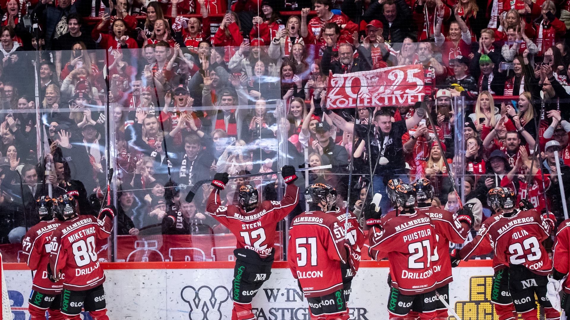 Ishockeyspelare i röda tröjor står längs sargen under en match med en full läktare av supportrar i bakgrunden.