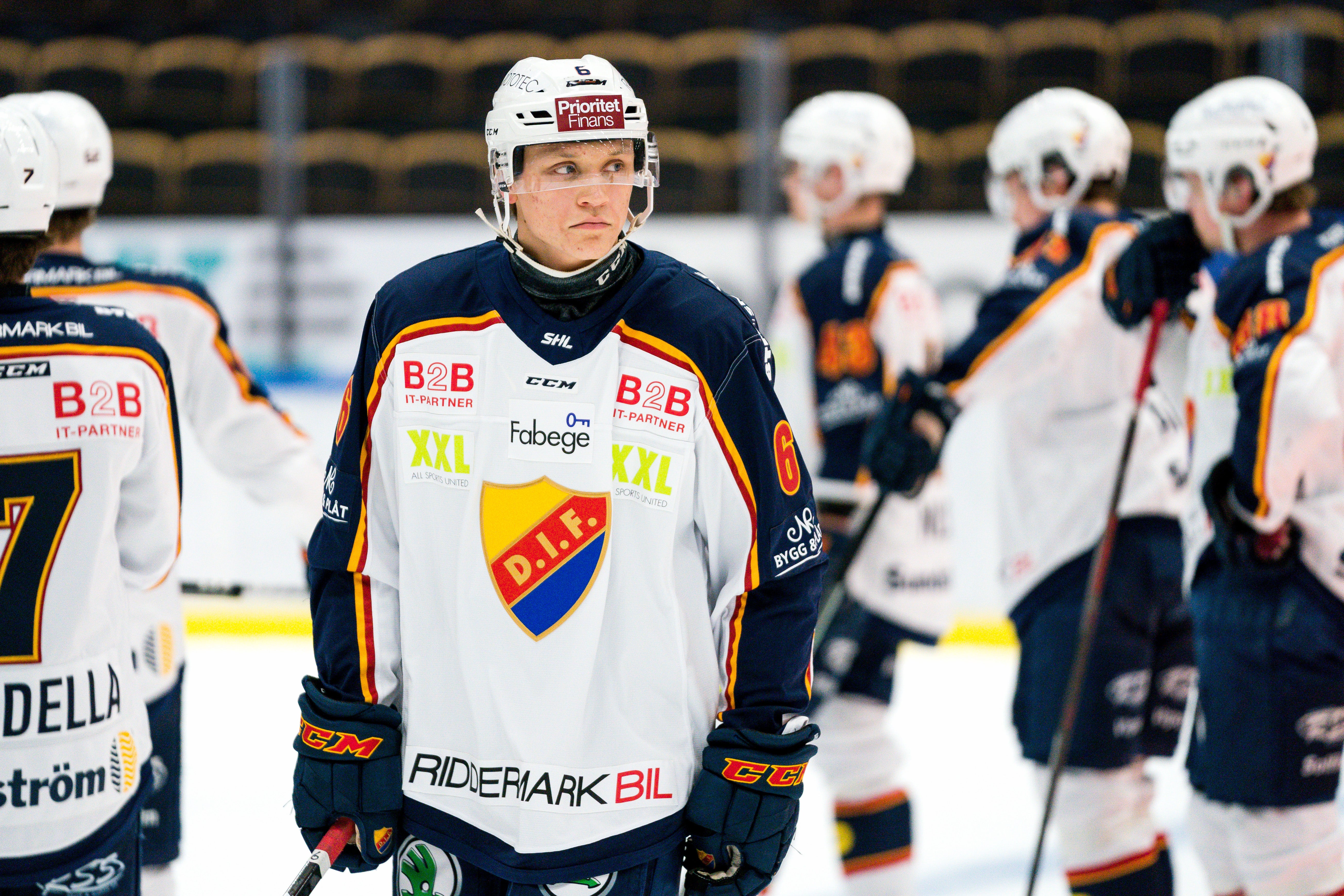 Ishockeyspelare i vit och marinblå matchdräkt står på isen med andra lagkamrater i bakgrunden under en ishockeymatch.
