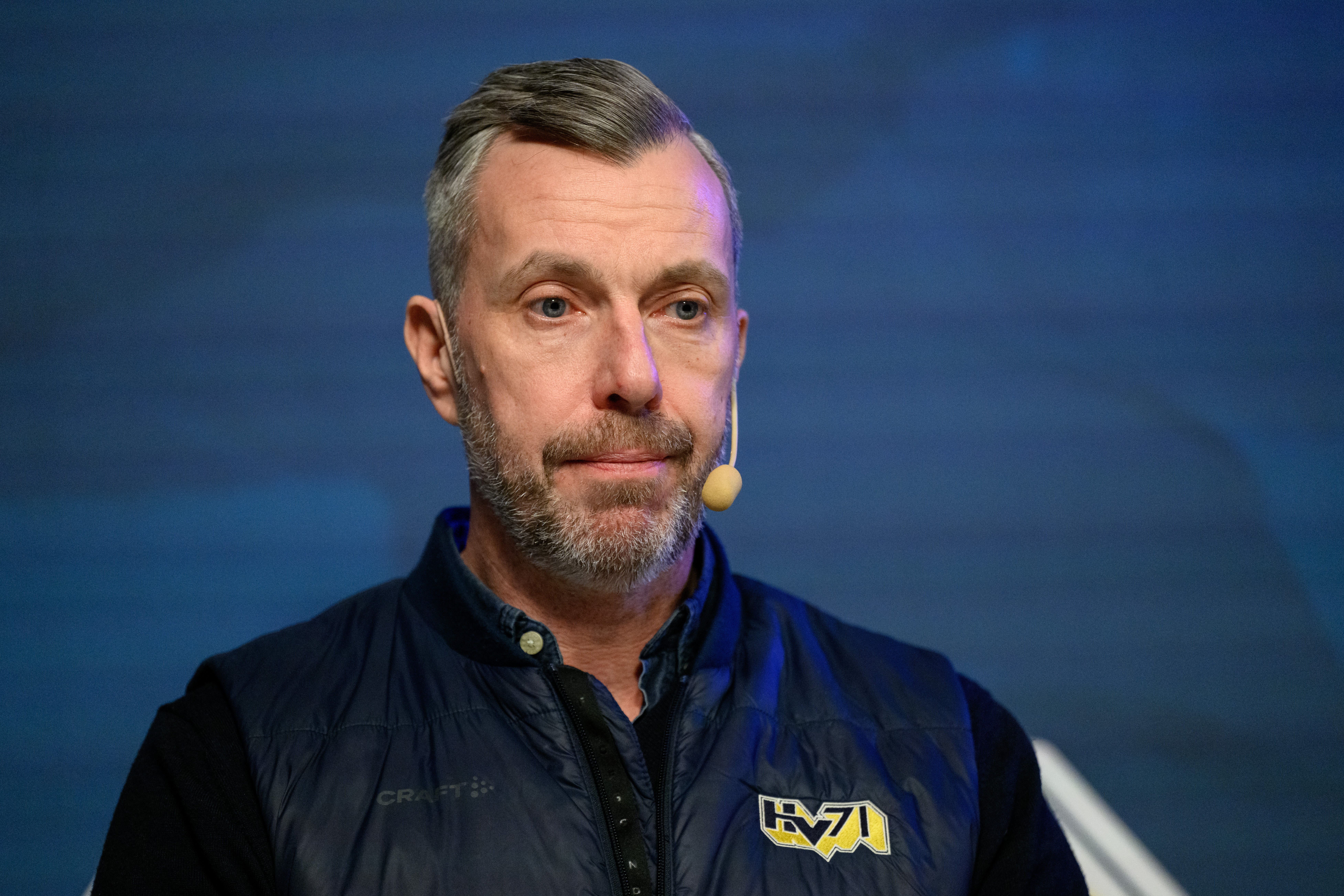 Porträtt av en man med skägg som bär en svart jacka med gul logga. Han är fotograferad mot en blå bakgrund och har ett headset-mikrofon på.