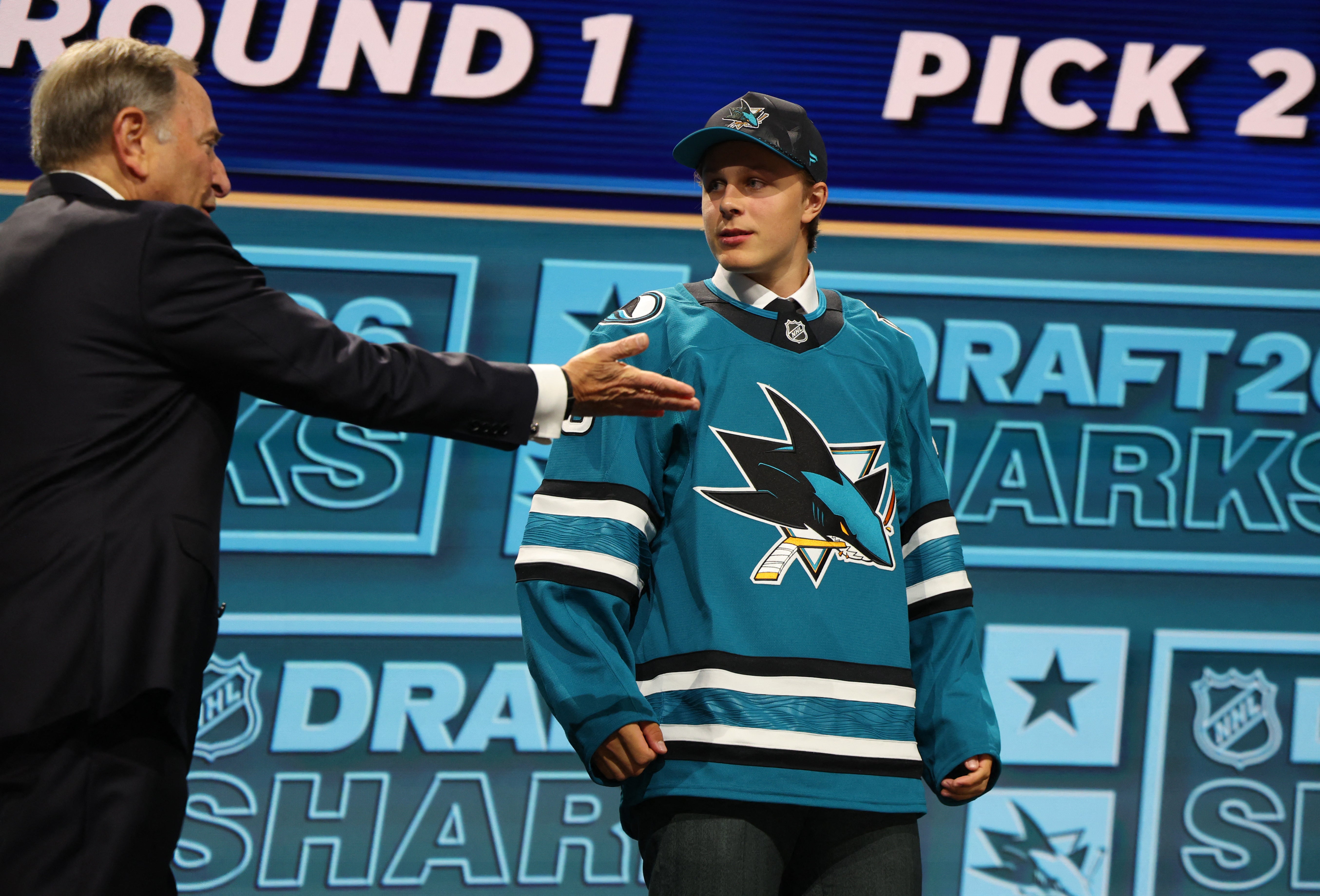En ung hockeyspelare i San Jose Sharks tröja presenteras på NHL-draften av en man i svart kostym på scenen.