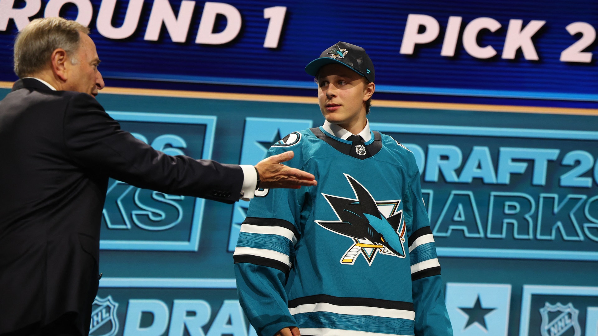 En ung hockeyspelare i San Jose Sharks tröja presenteras på NHL-draften av en man i svart kostym på scenen.