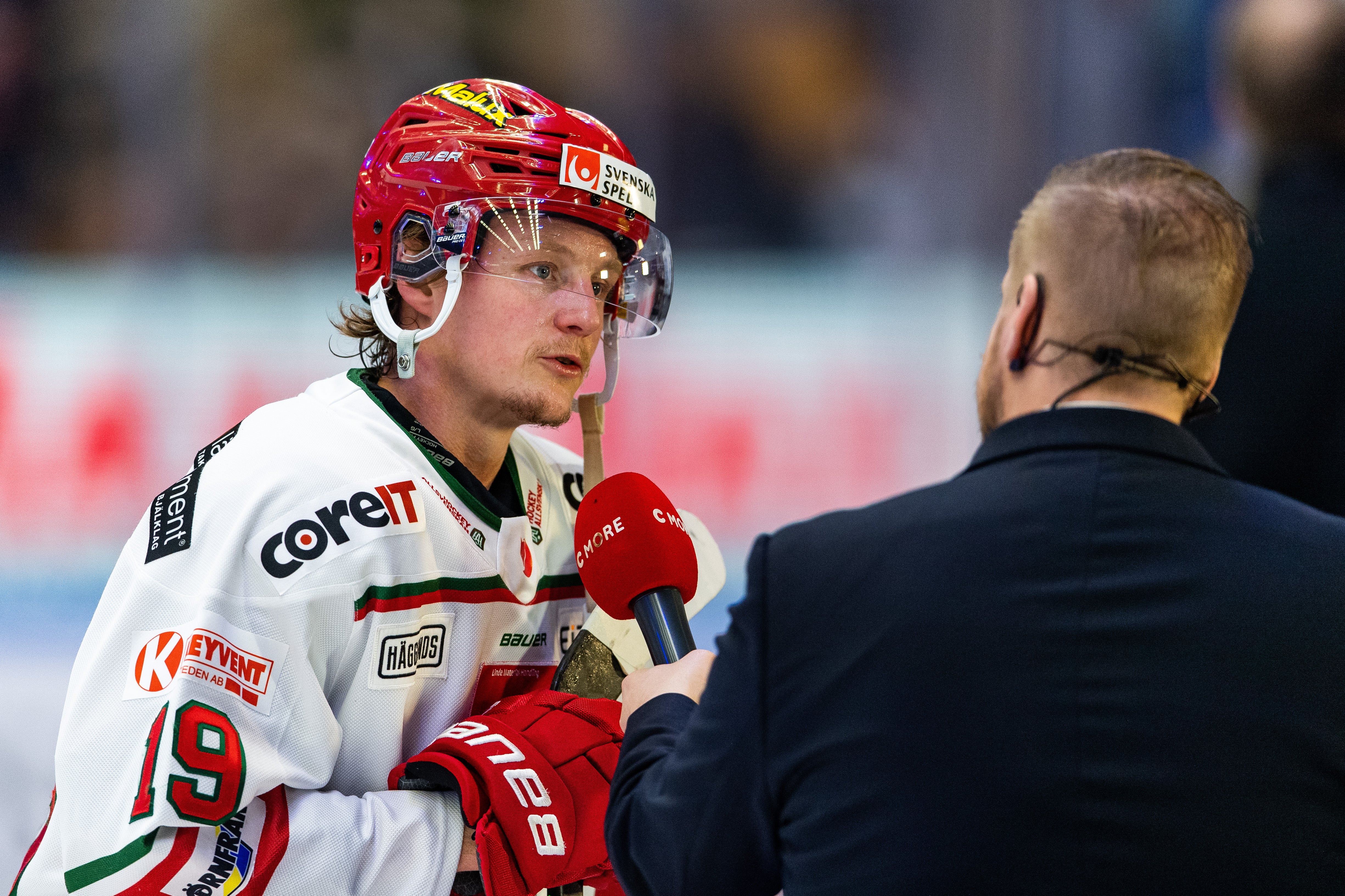 En ishockeyspelare i vit tröja och röd hjälm intervjuas av en reporter med mikrofon.