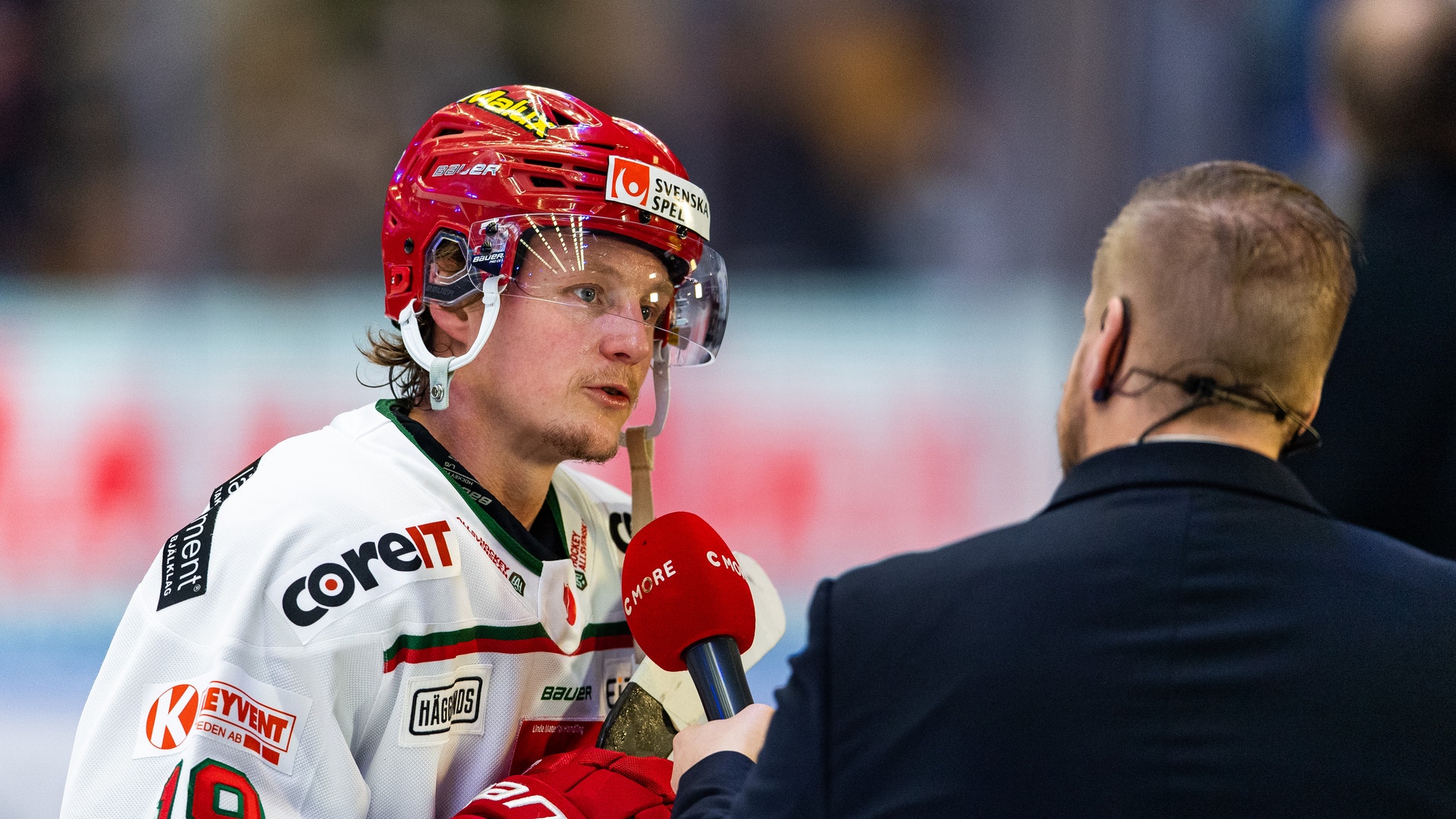 En ishockeyspelare i vit tröja och röd hjälm intervjuas av en reporter med mikrofon.