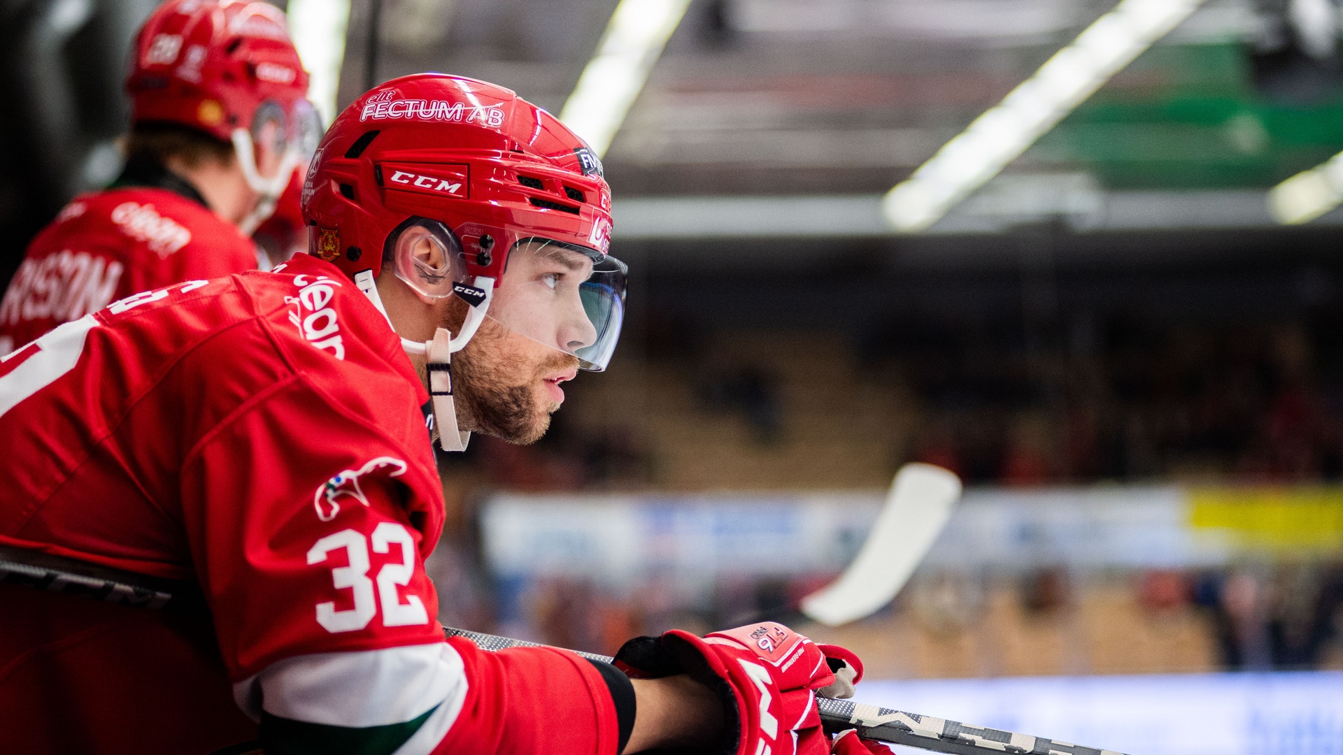 Ishockeyspelare i röd dräkt med nummer 32 fokuserad på spelet, bärande röd hjälm och glasögon med ishockeyklubba i hand.