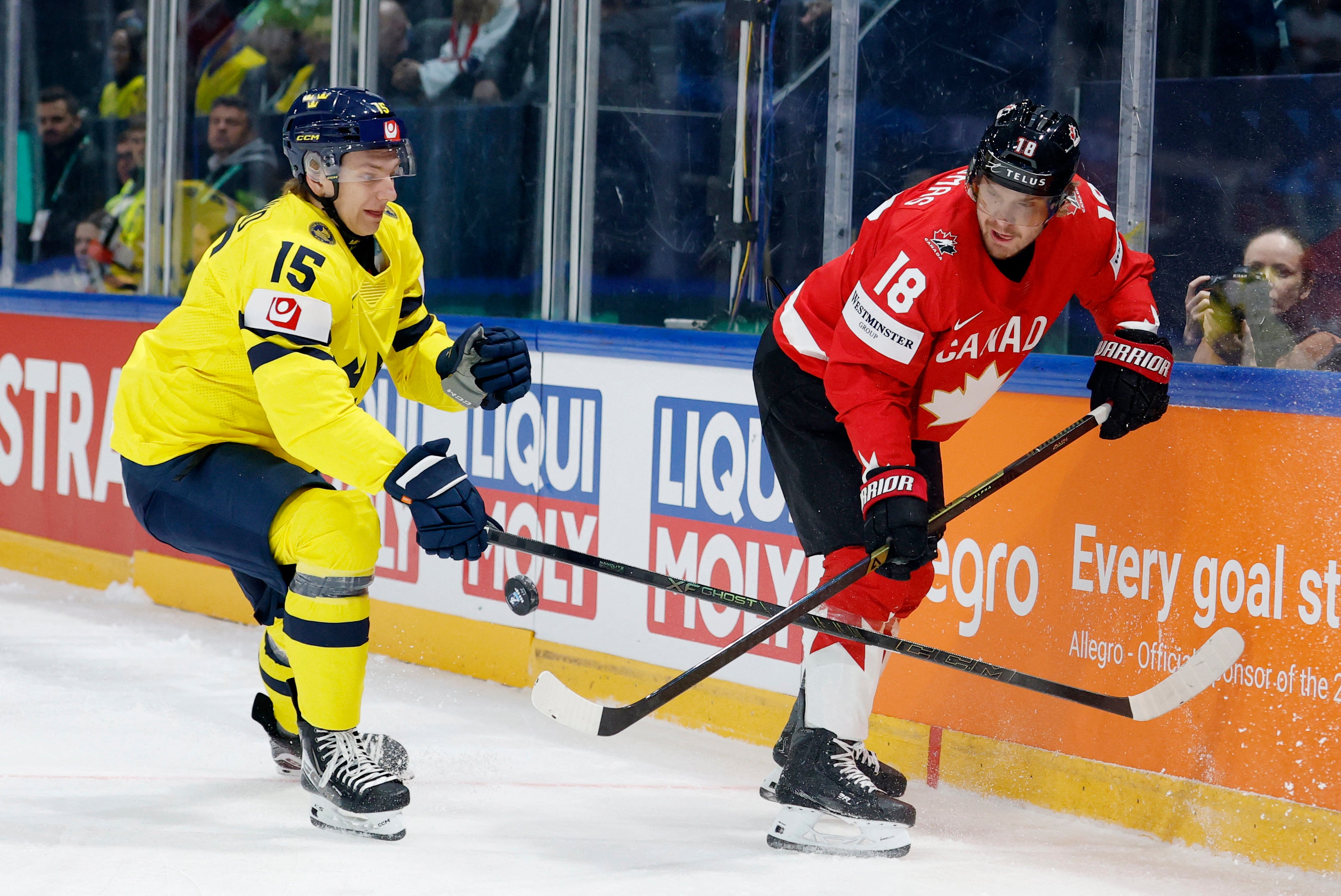 Två ishockeyspelare under en match. En spelare i gul tröja och en spelare i röd tröja med Kanadas flagga kämpar om pucken på isen. Publiken syns i bakgrunden bakom glaset.