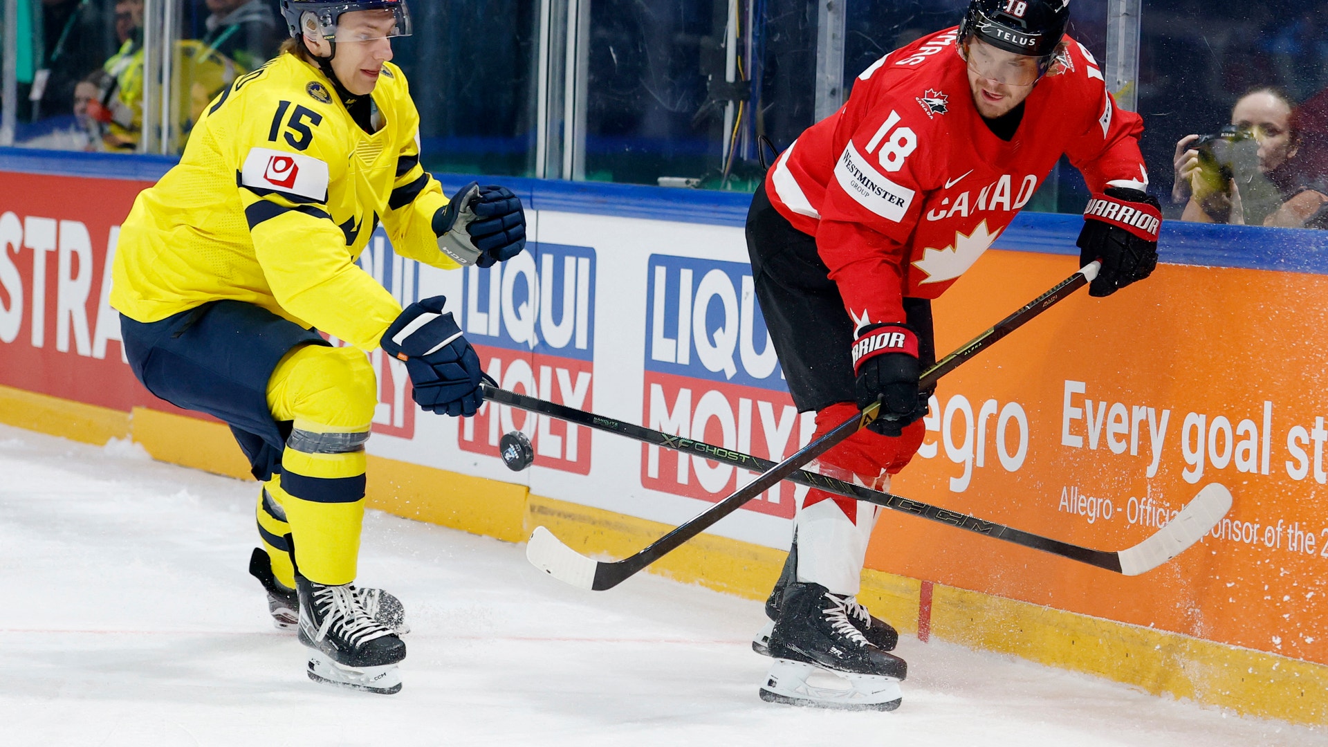 Två ishockeyspelare under en match. En spelare i gul tröja och en spelare i röd tröja med Kanadas flagga kämpar om pucken på isen. Publiken syns i bakgrunden bakom glaset.