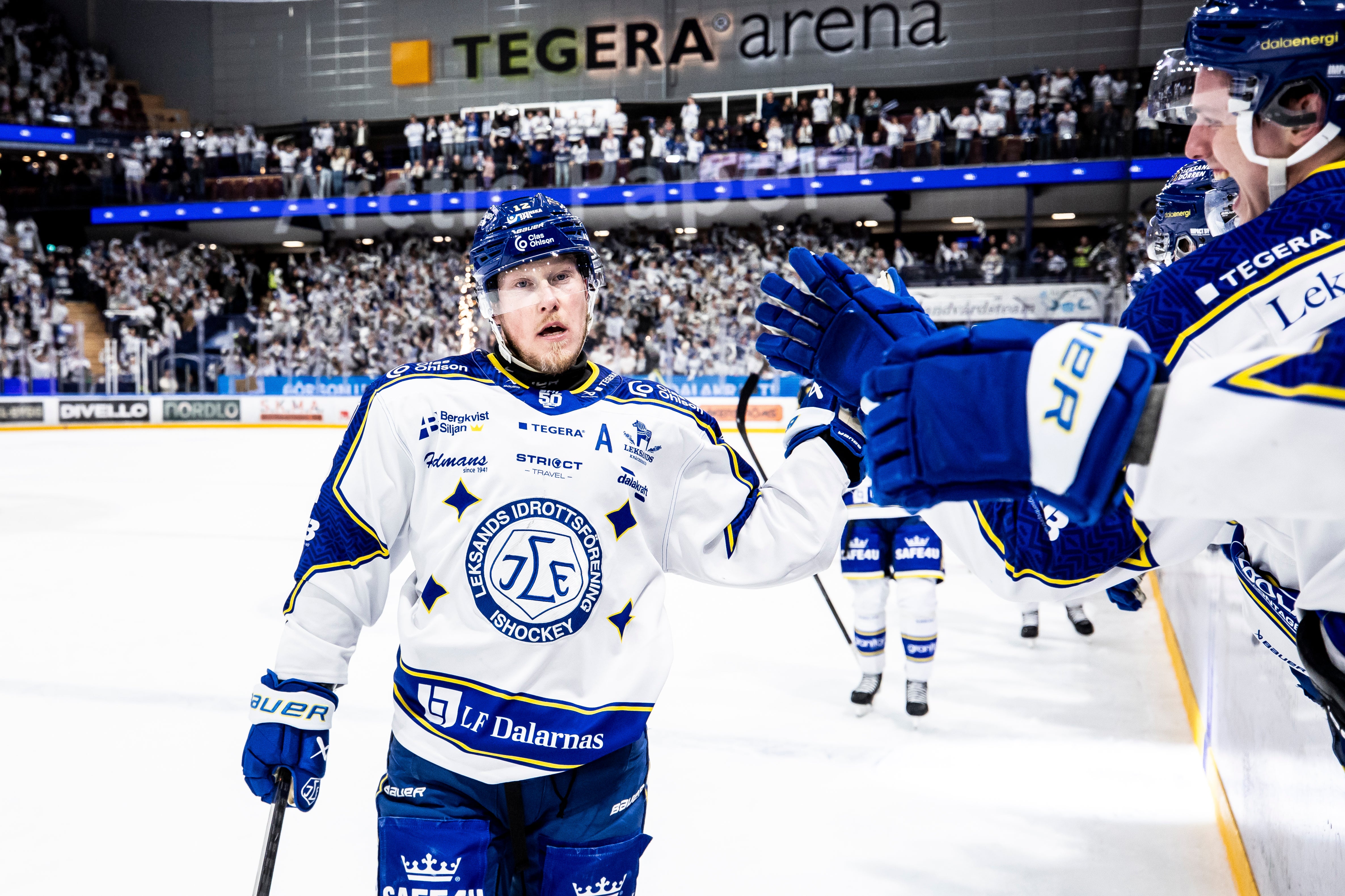 Ishockeyspelare i blå och vit tröja under en match på Tegera Arena. Spelaren står på isen med sin hockeyklubba och publik syns i bakgrunden på läktaren.