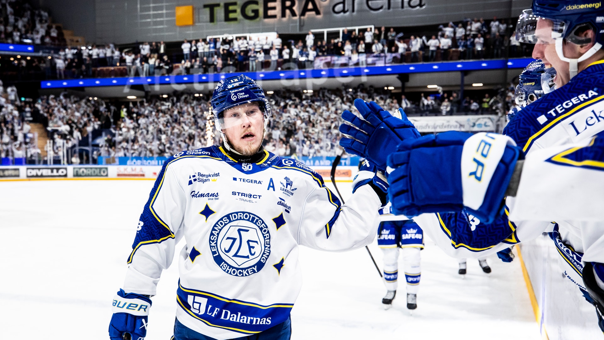Ishockeyspelare i blå och vit tröja under en match på Tegera Arena. Spelaren står på isen med sin hockeyklubba och publik syns i bakgrunden på läktaren.