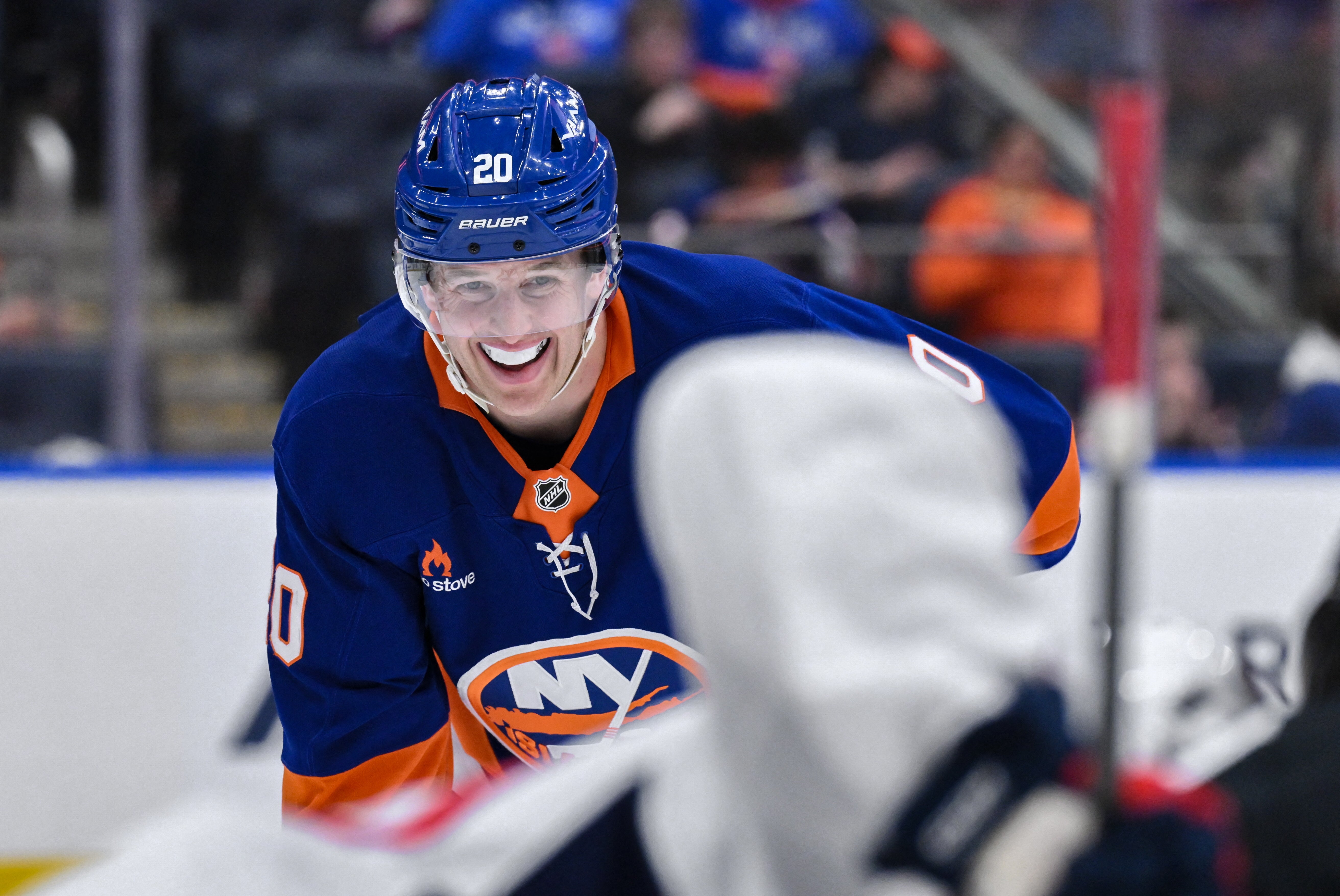 En hockeyspelare i blå och orange Islanders-uniform ler mot kameran under en match. Spelaren bär nummer 20 och en blå hjälm.