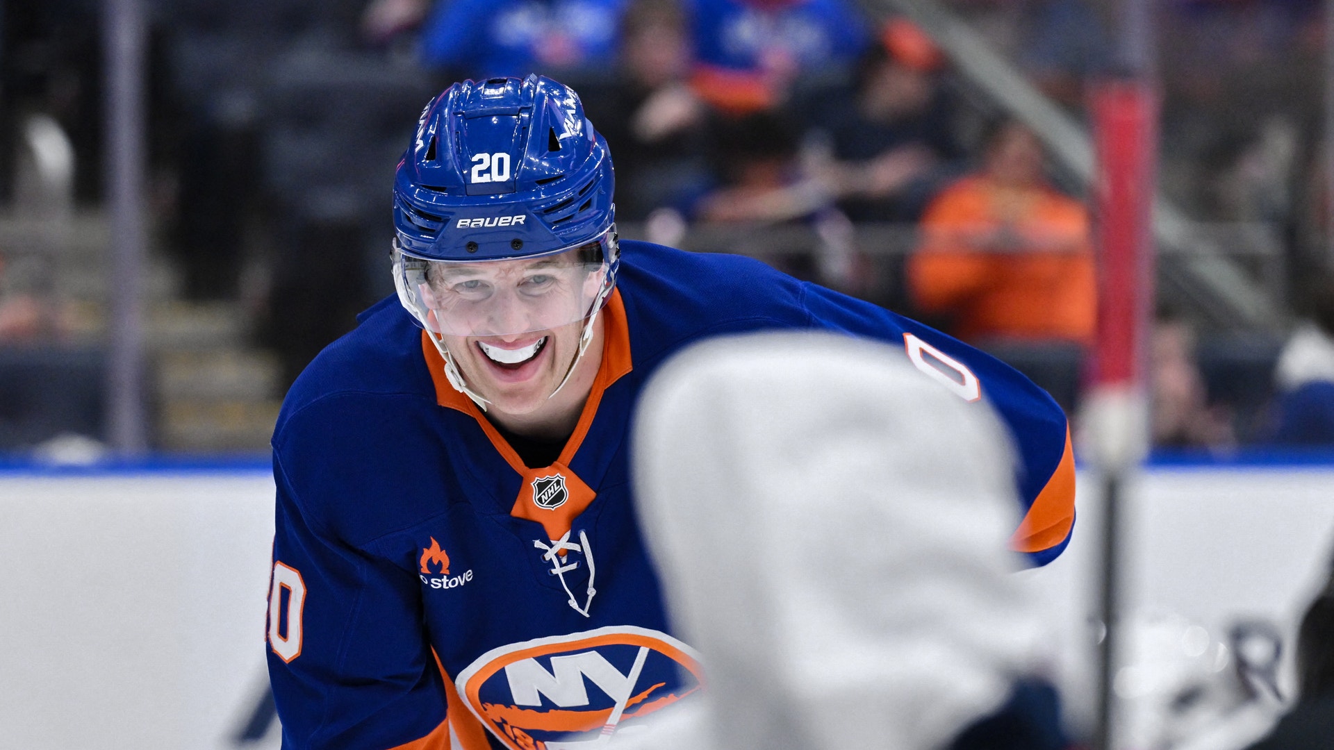 En hockeyspelare i blå och orange Islanders-uniform ler mot kameran under en match. Spelaren bär nummer 20 och en blå hjälm.