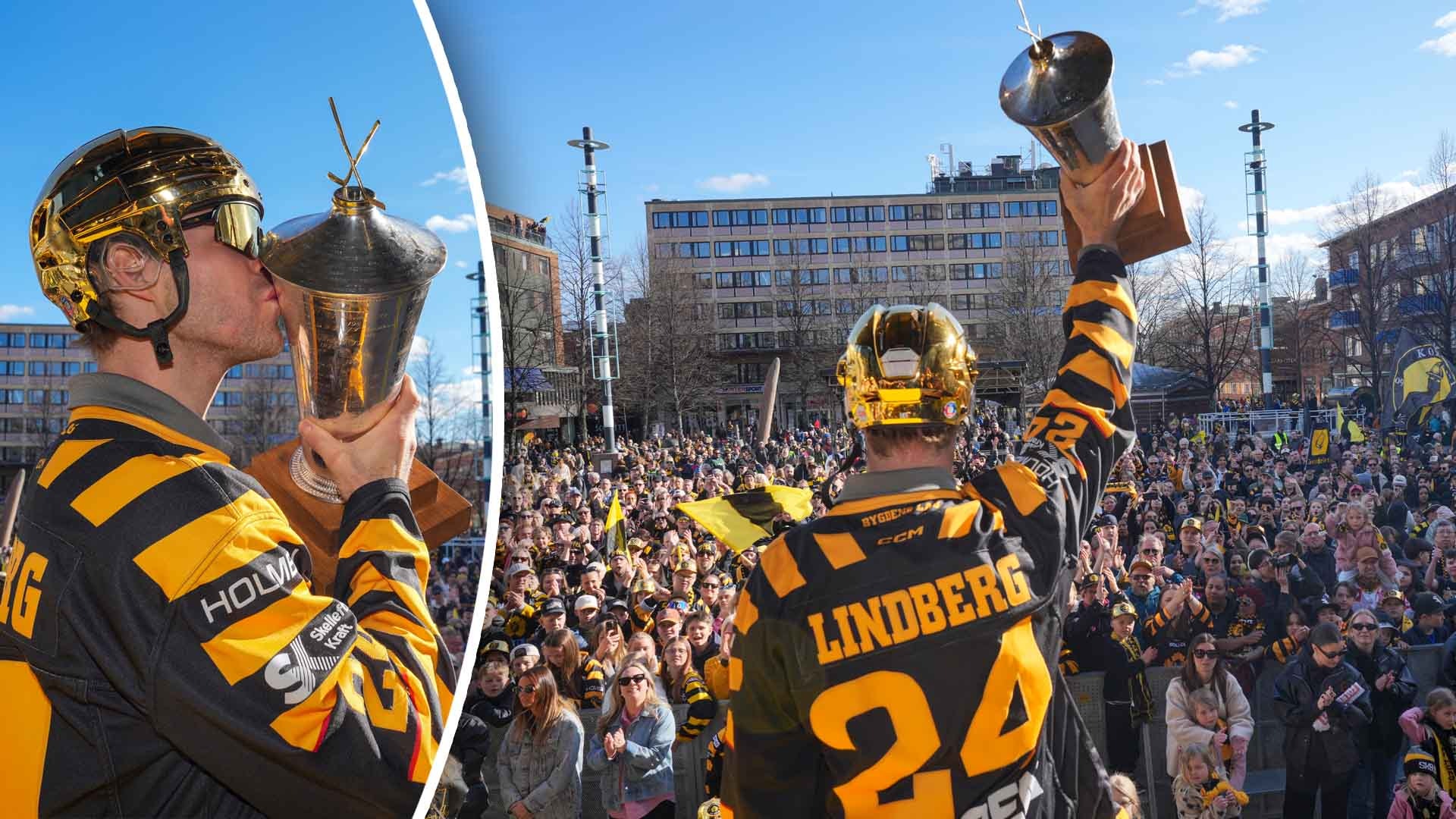 Hockeyspelare i gul och svart uniform firar med en stor pokal framför en stor folksamling på en öppen plats i stadsmiljö.