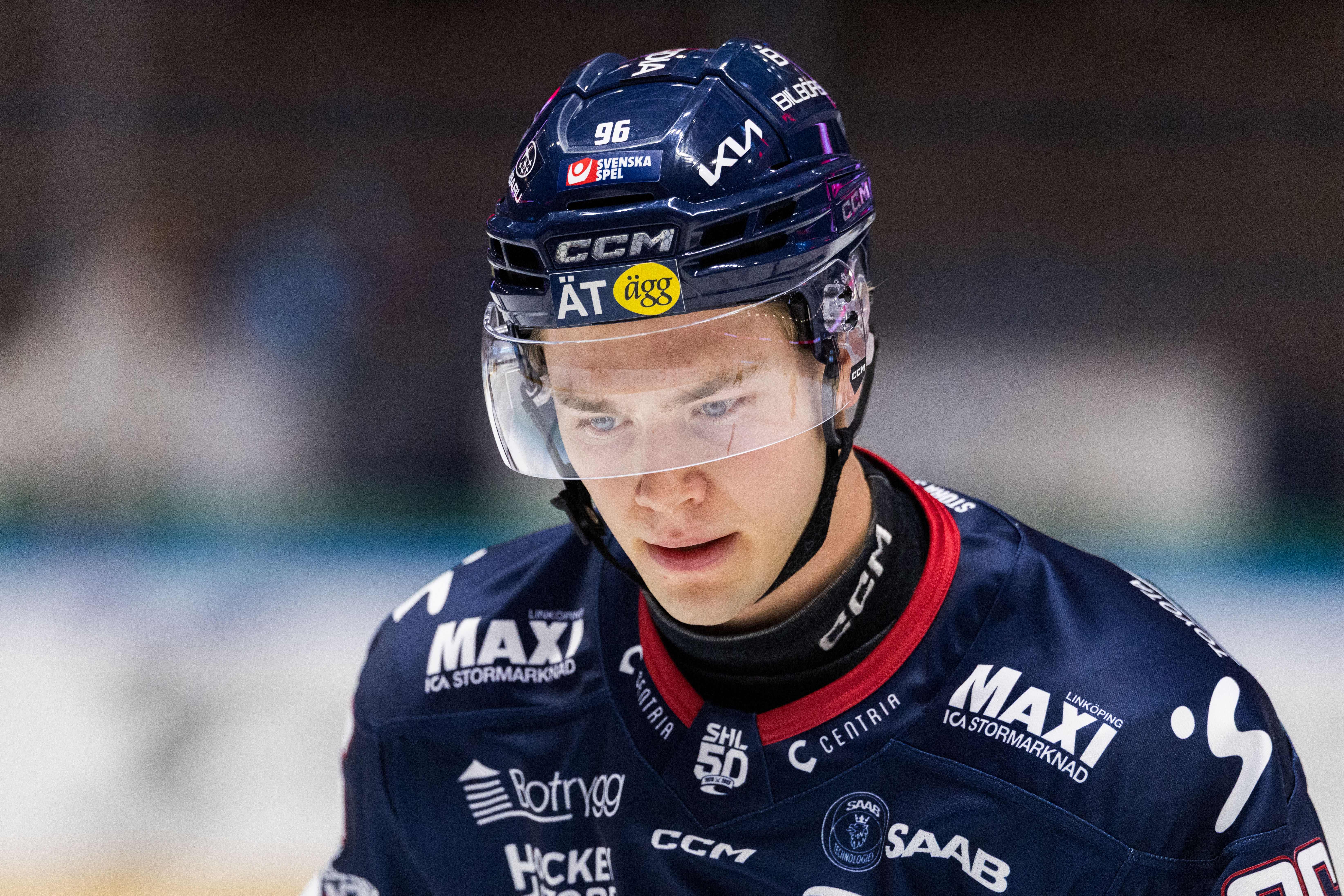 Ishockeyspelare i mörkblå tröja med sponsorlogotyper under en match. Spelaren bär mörkblå hjälm med skyddsglas och är fokuserad på spelet.