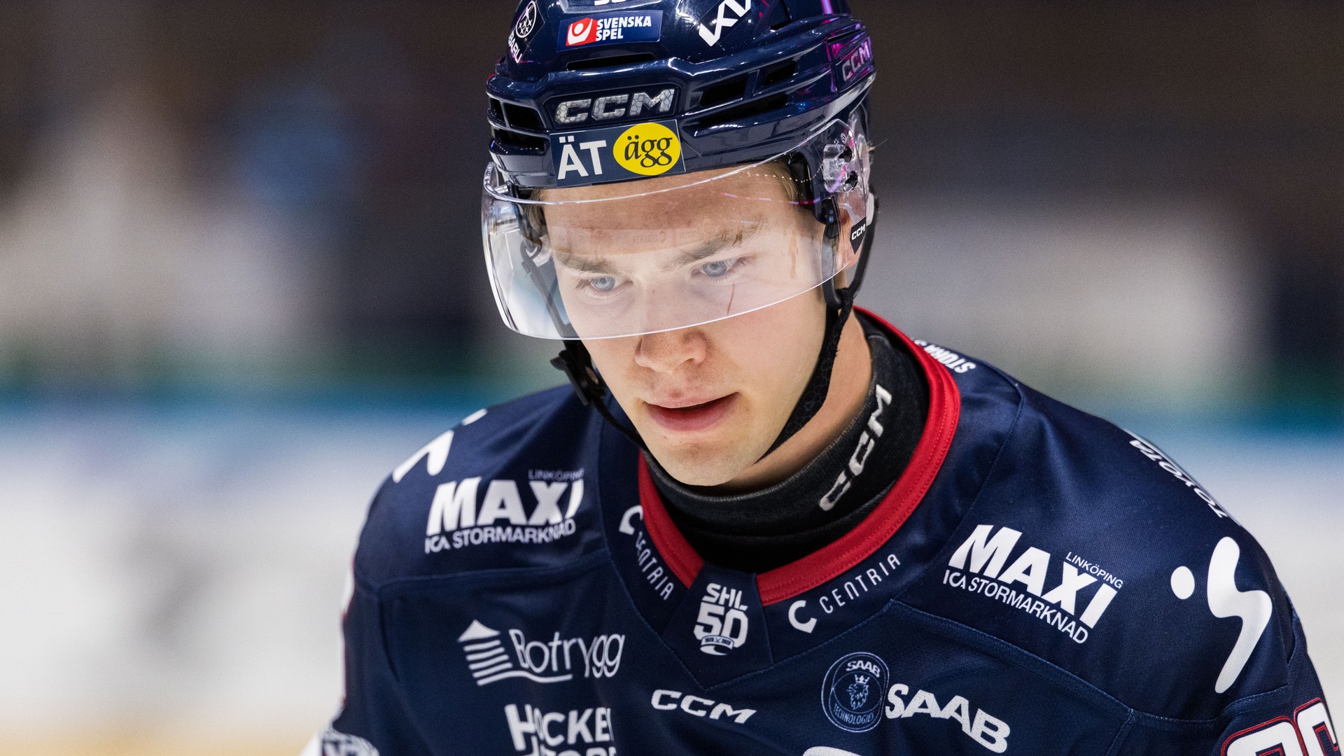 Ishockeyspelare i mörkblå tröja med sponsorlogotyper under en match. Spelaren bär mörkblå hjälm med skyddsglas och är fokuserad på spelet.