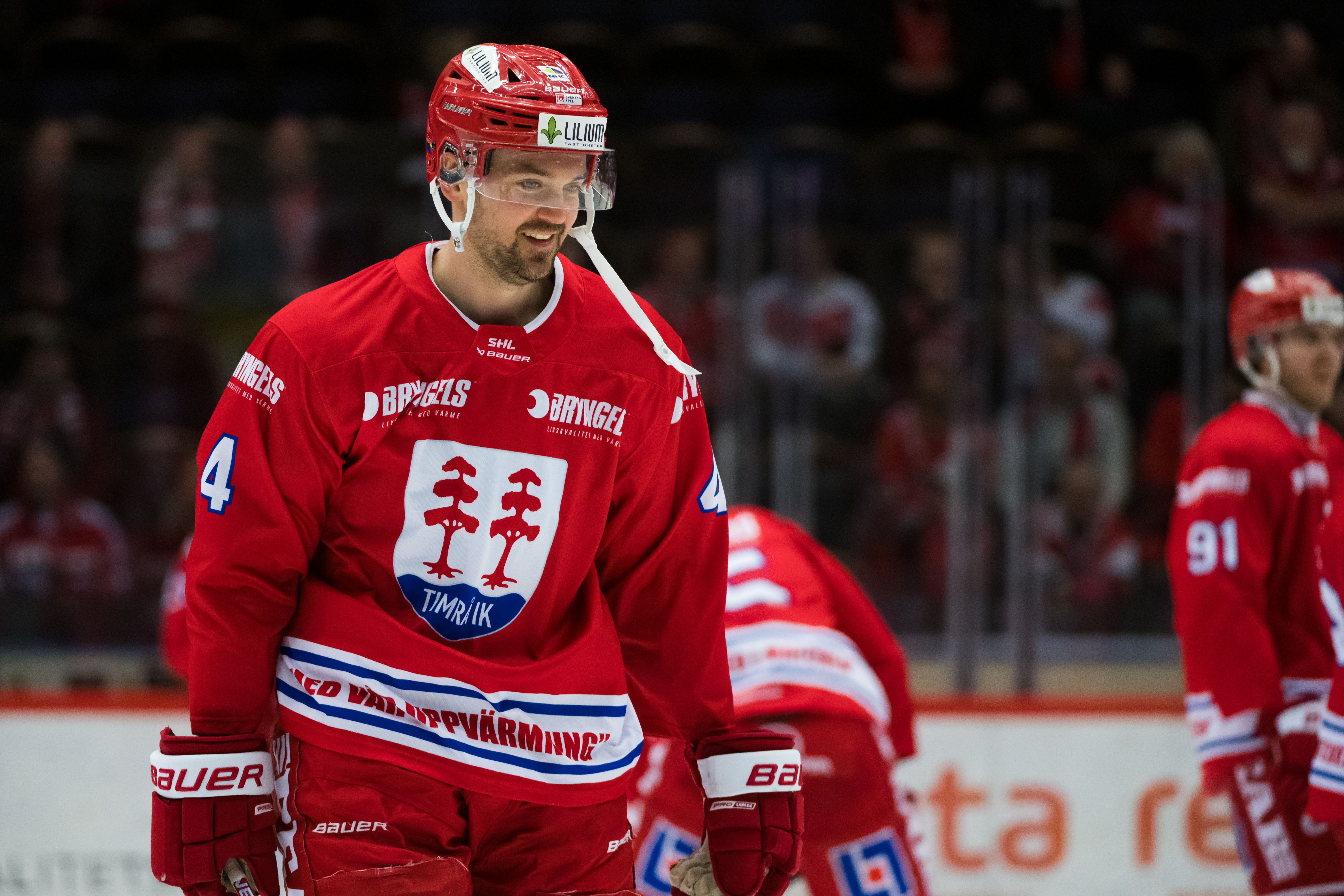 Ishockeyspelare i röd landslagströja under en match. Spelaren bär nummer 4 och en röd hjälm, och står på isen med publik synlig i bakgrunden.