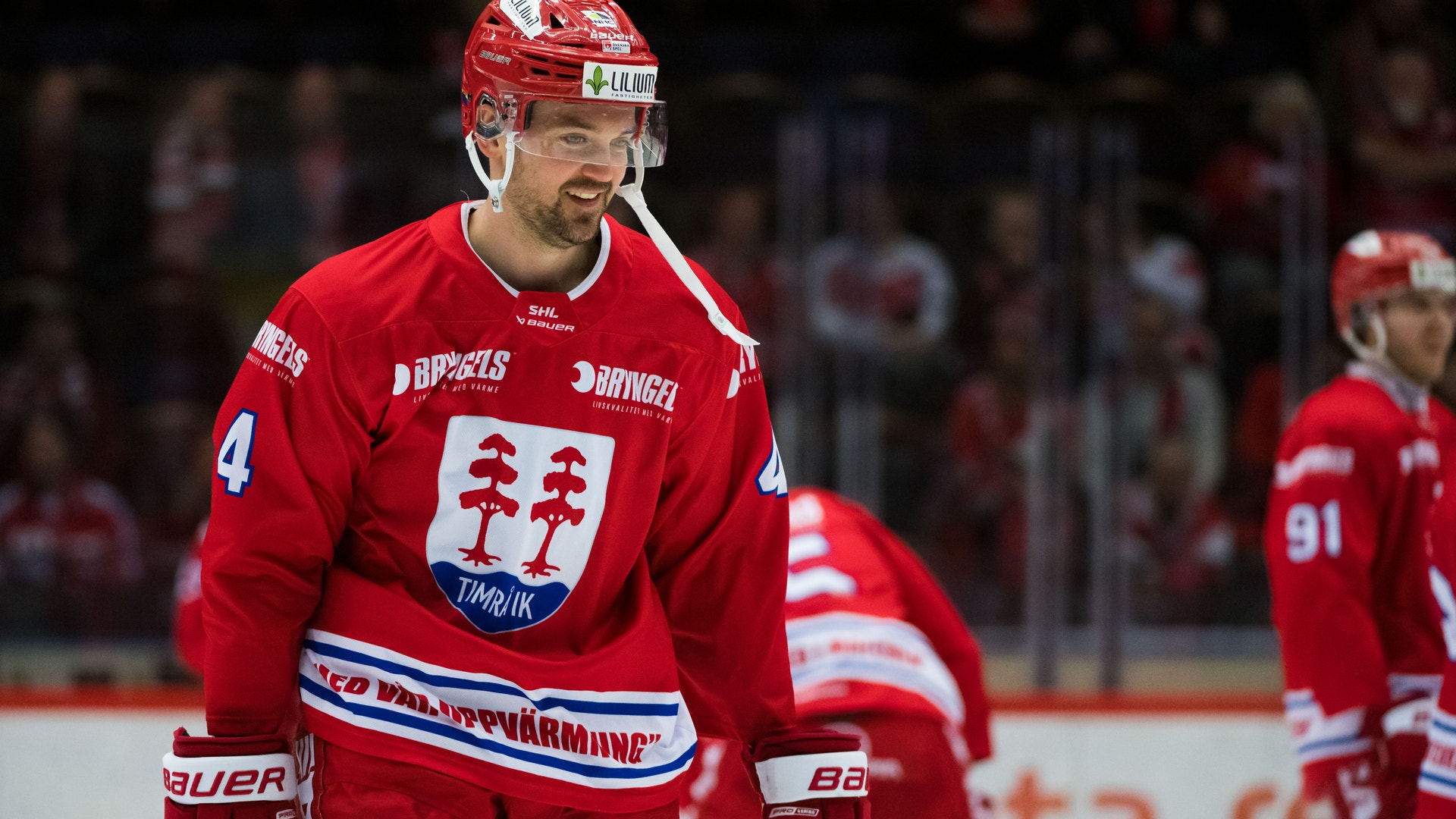 Ishockeyspelare i röd landslagströja under en match. Spelaren bär nummer 4 och en röd hjälm, och står på isen med publik synlig i bakgrunden.