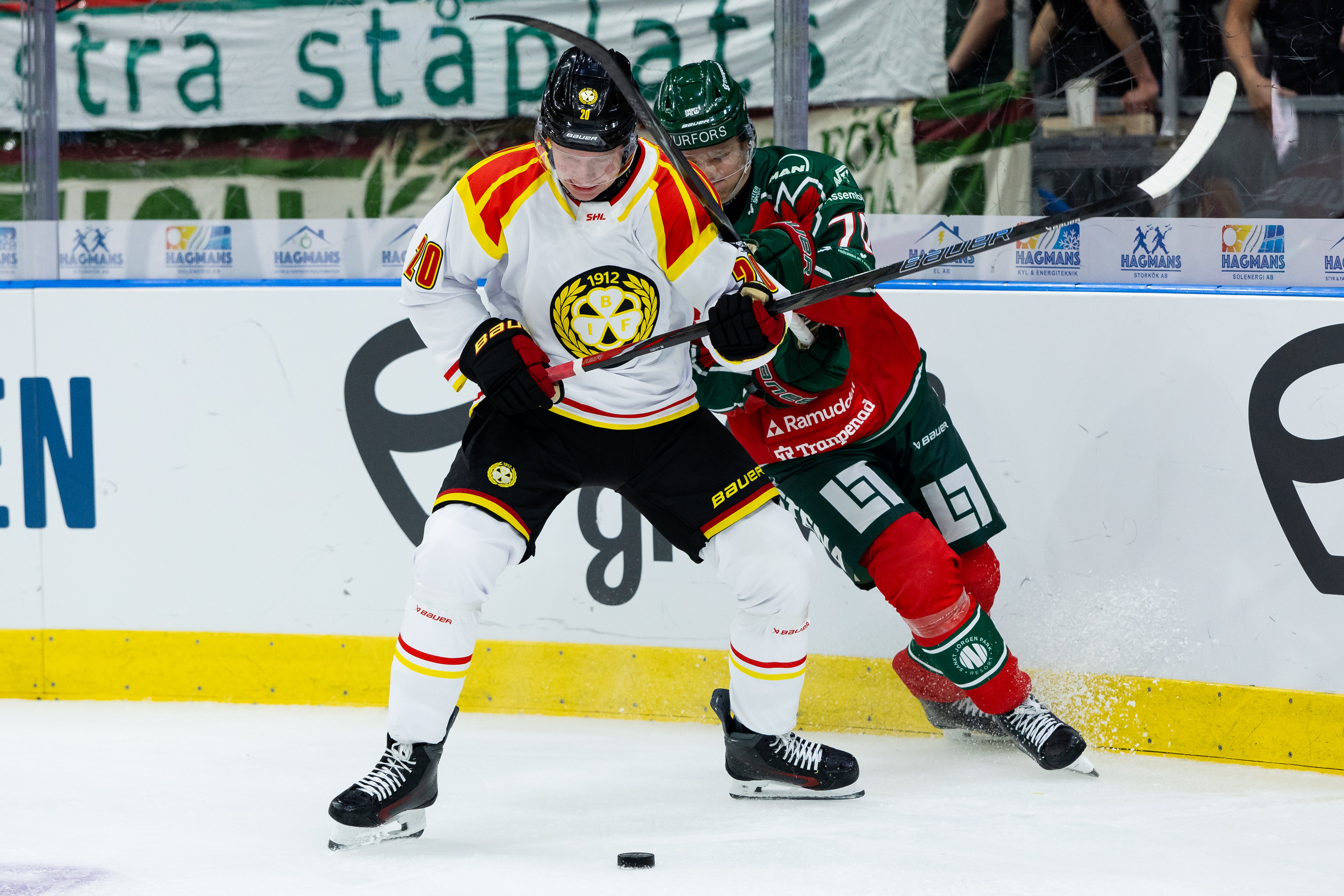 Två ishockeyspelare i kamp om pucken vid sargen under en match. Spelaren i vit och gul tröja trycker mot en motståndare i grön och röd dräkt.