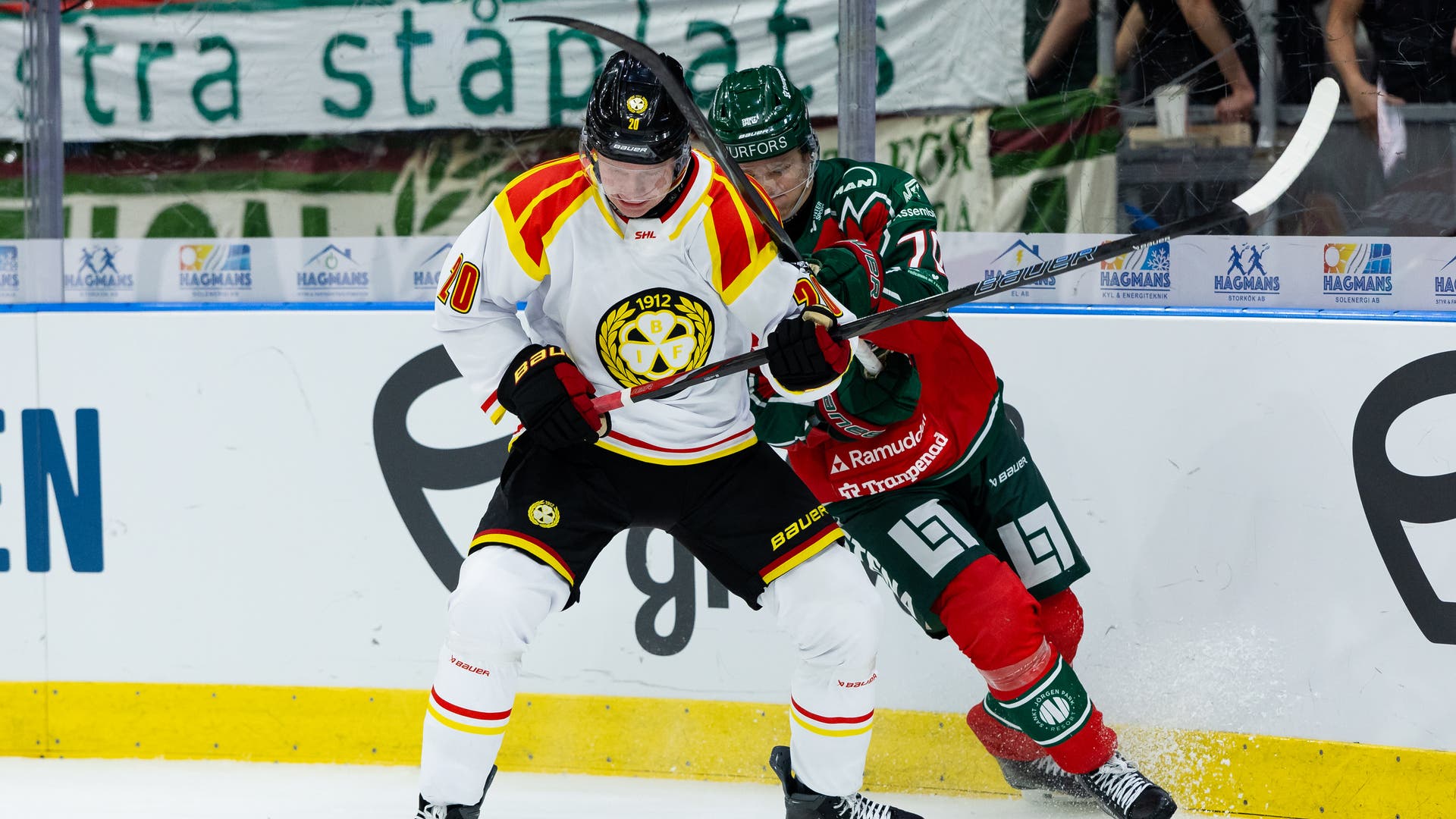 Två ishockeyspelare i kamp om pucken vid sargen under en match. Spelaren i vit och gul tröja trycker mot en motståndare i grön och röd dräkt.