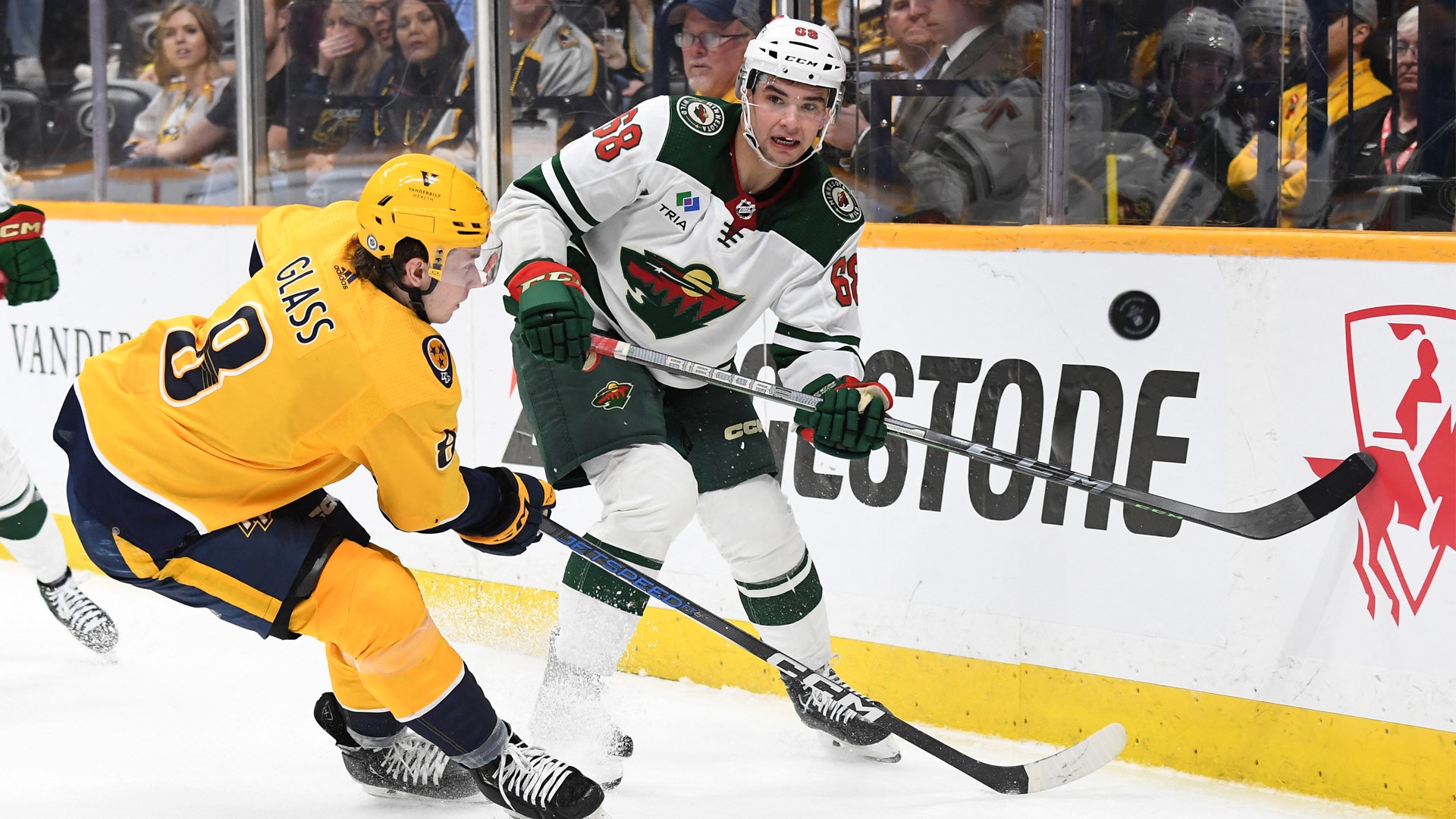 Två ishockeyspelare kämpar om pucken längs banden under en match. En spelare är klädd i gul tröja och den andra i vit och svart tröja.