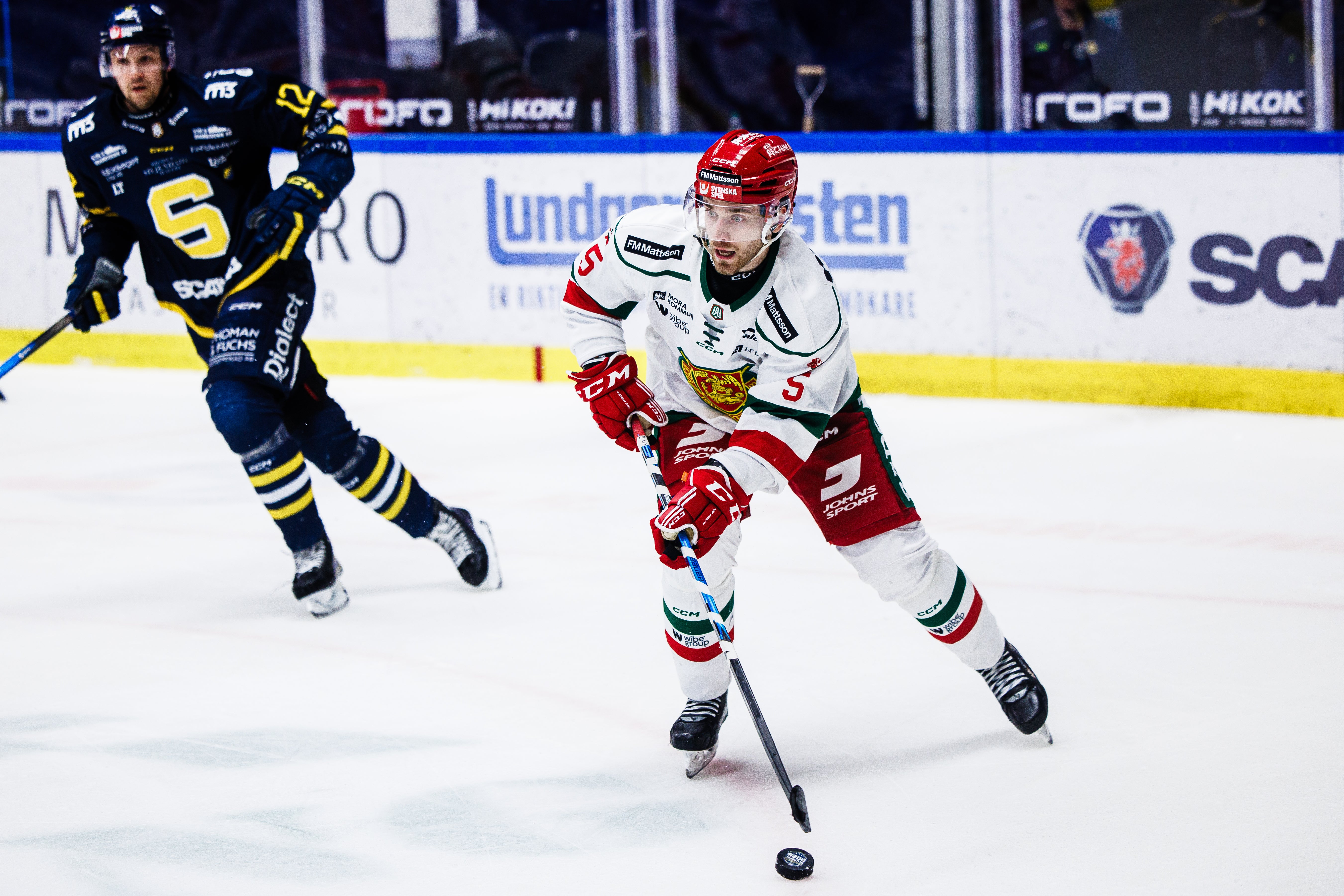 Ishockeymatch där en spelare i röd och vit tröja dribblar med pucken medan en motspelare i mörkblå och gul tröja försöker försvara på isen.