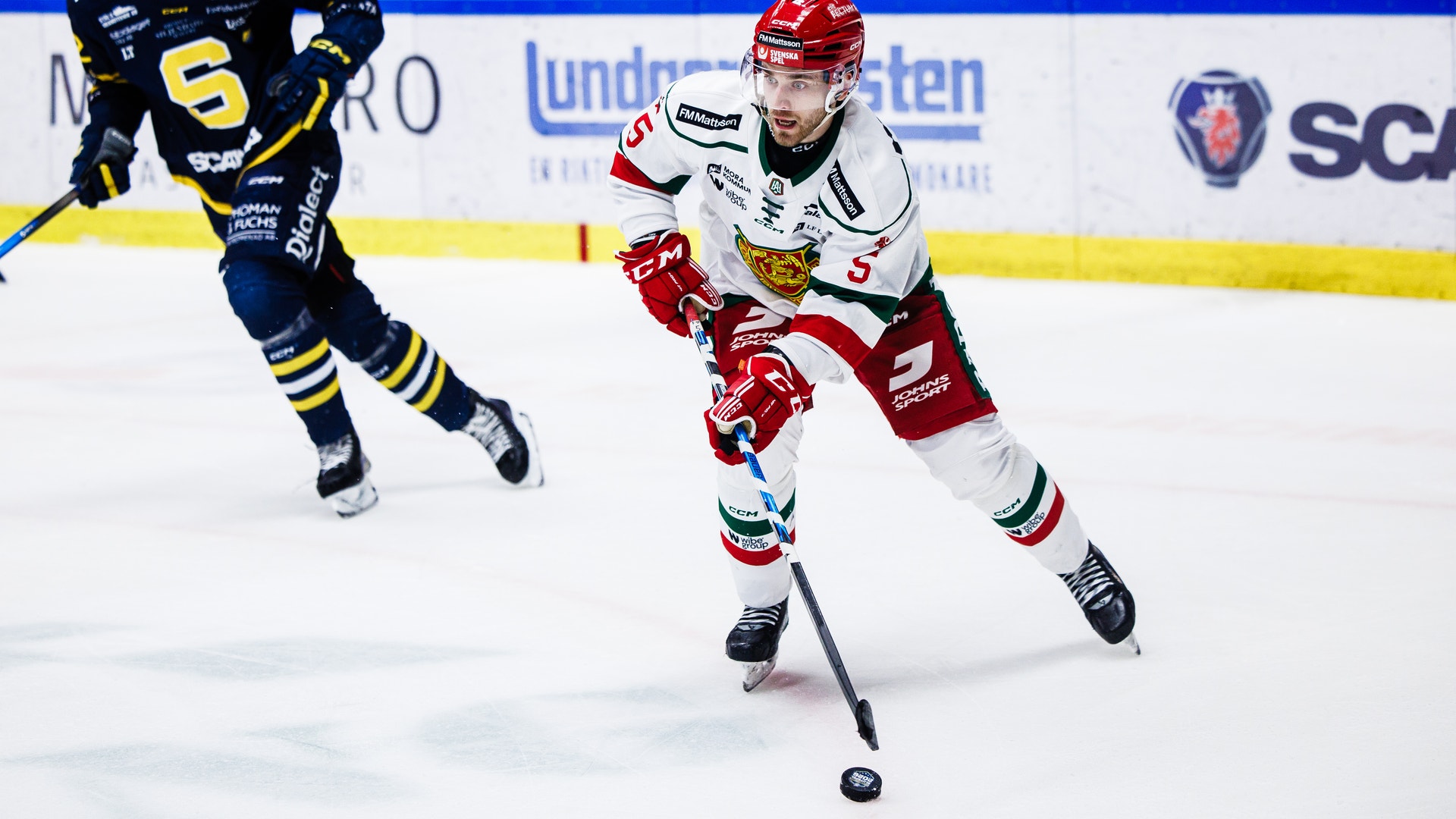 Ishockeymatch där en spelare i röd och vit tröja dribblar med pucken medan en motspelare i mörkblå och gul tröja försöker försvara på isen.