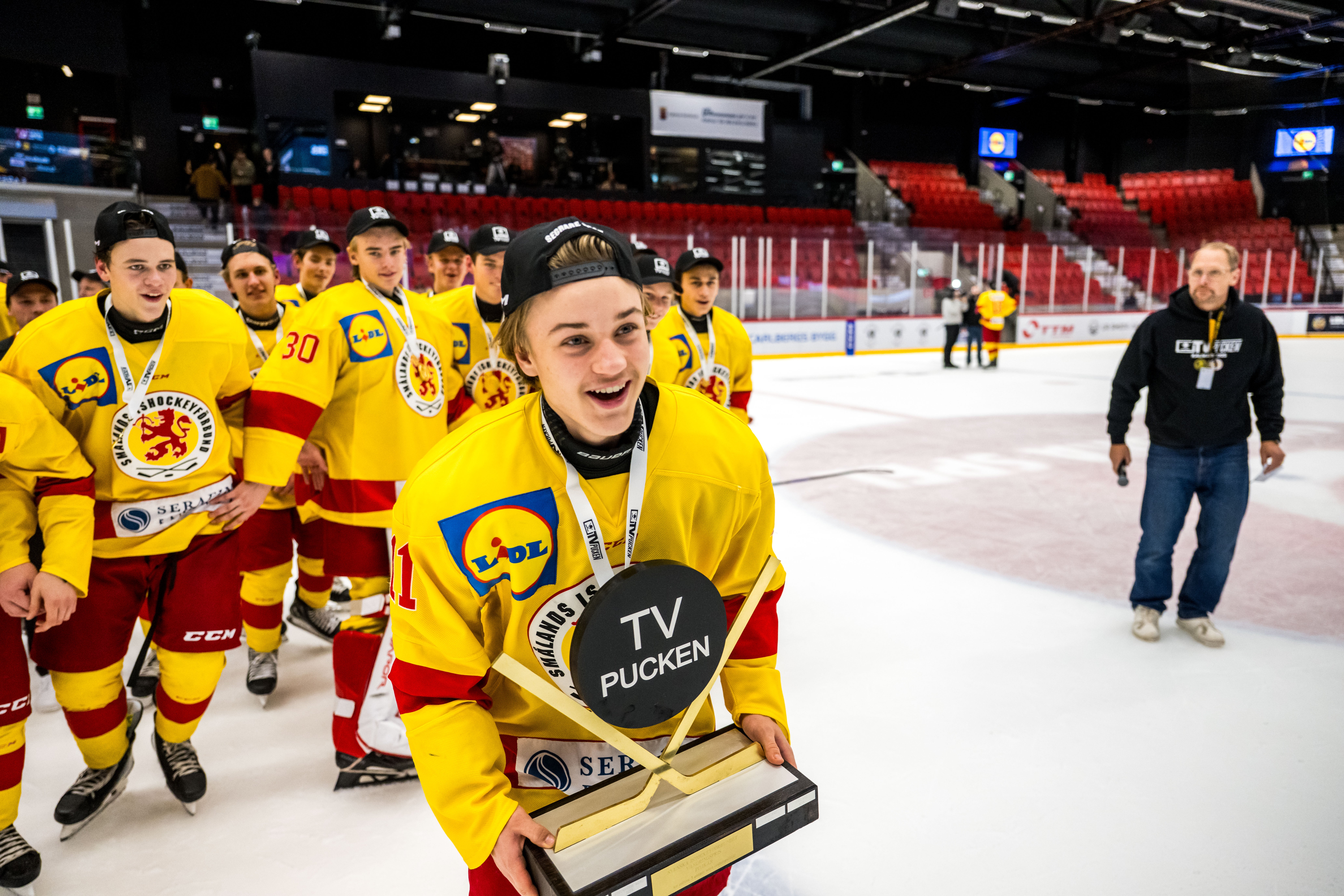 Hockeyspelare i gul och röd tröja håller i en pokal på isen, omgiven av lagkamrater i samma färger.
