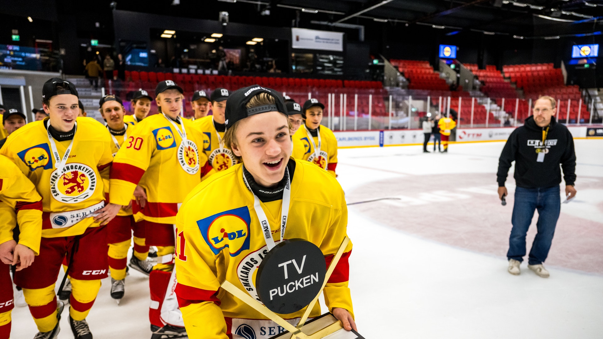 Hockeyspelare i gul och röd tröja håller i en pokal på isen, omgiven av lagkamrater i samma färger.