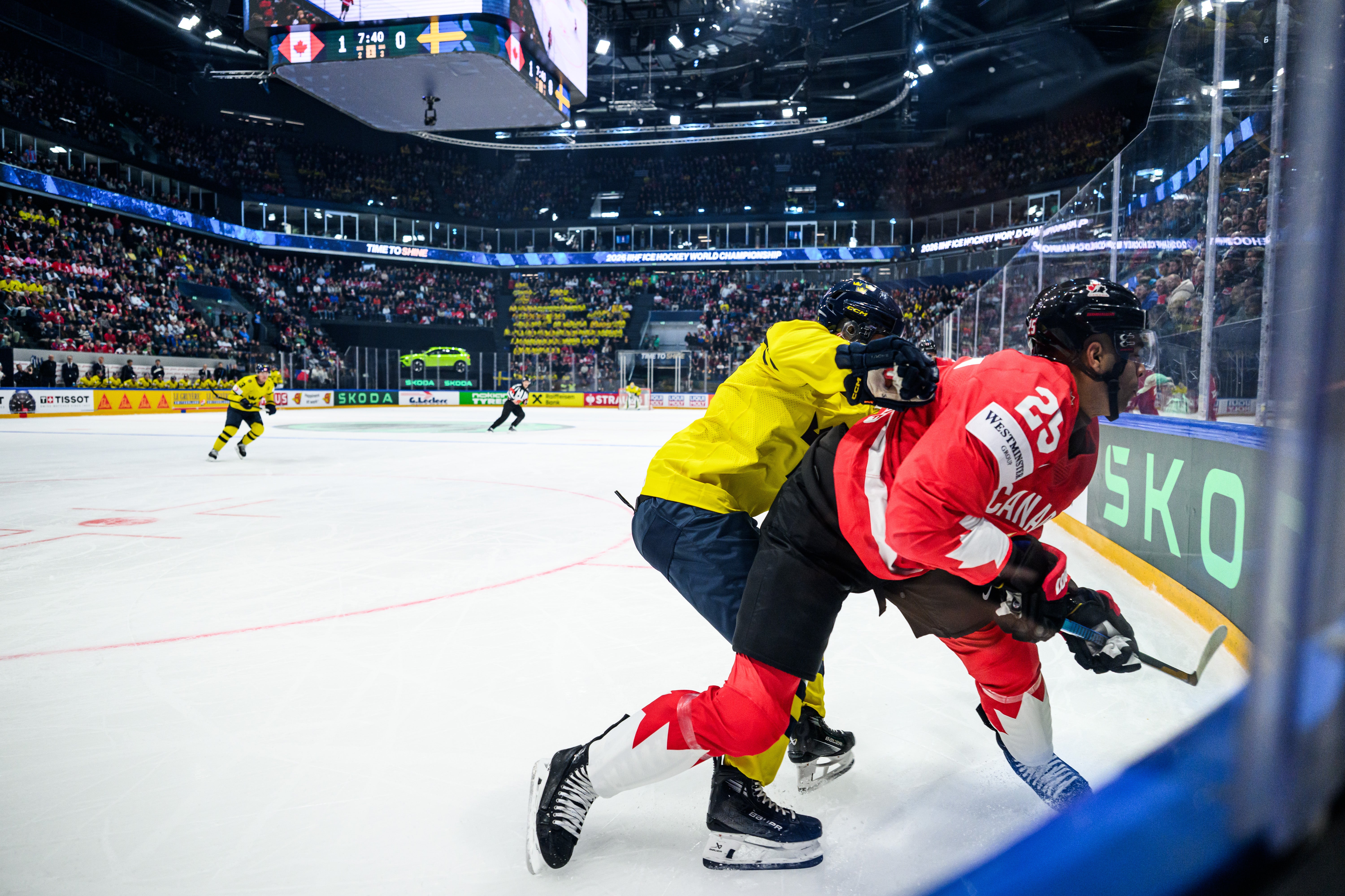 Två ishockeyspelare i kamp om pucken under en match i en fullsatt arena. En spelare i gul och svart uniform och en i röd uniform kämpar nära sargen.