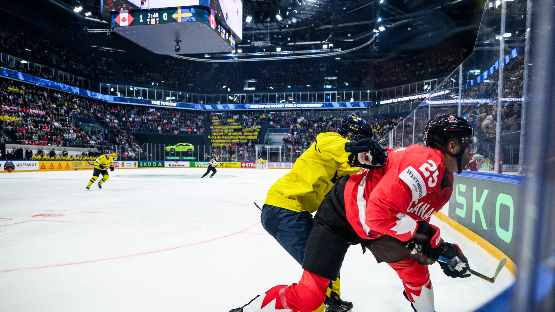 Två ishockeyspelare i kamp om pucken under en match i en fullsatt arena. En spelare i gul och svart uniform och en i röd uniform kämpar nära sargen.