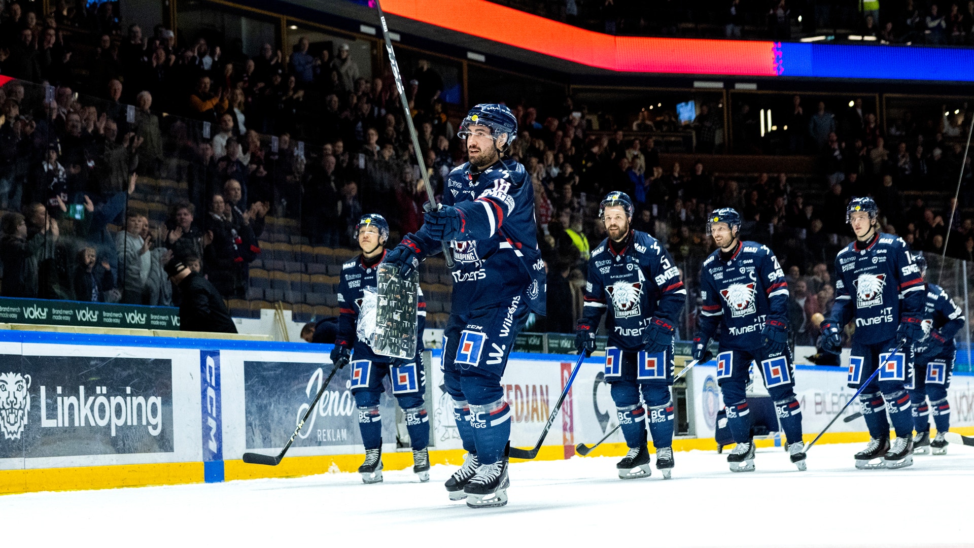Ishockeymatch med spelare i blåa tröjor på isen framför en fullsatt läktare.