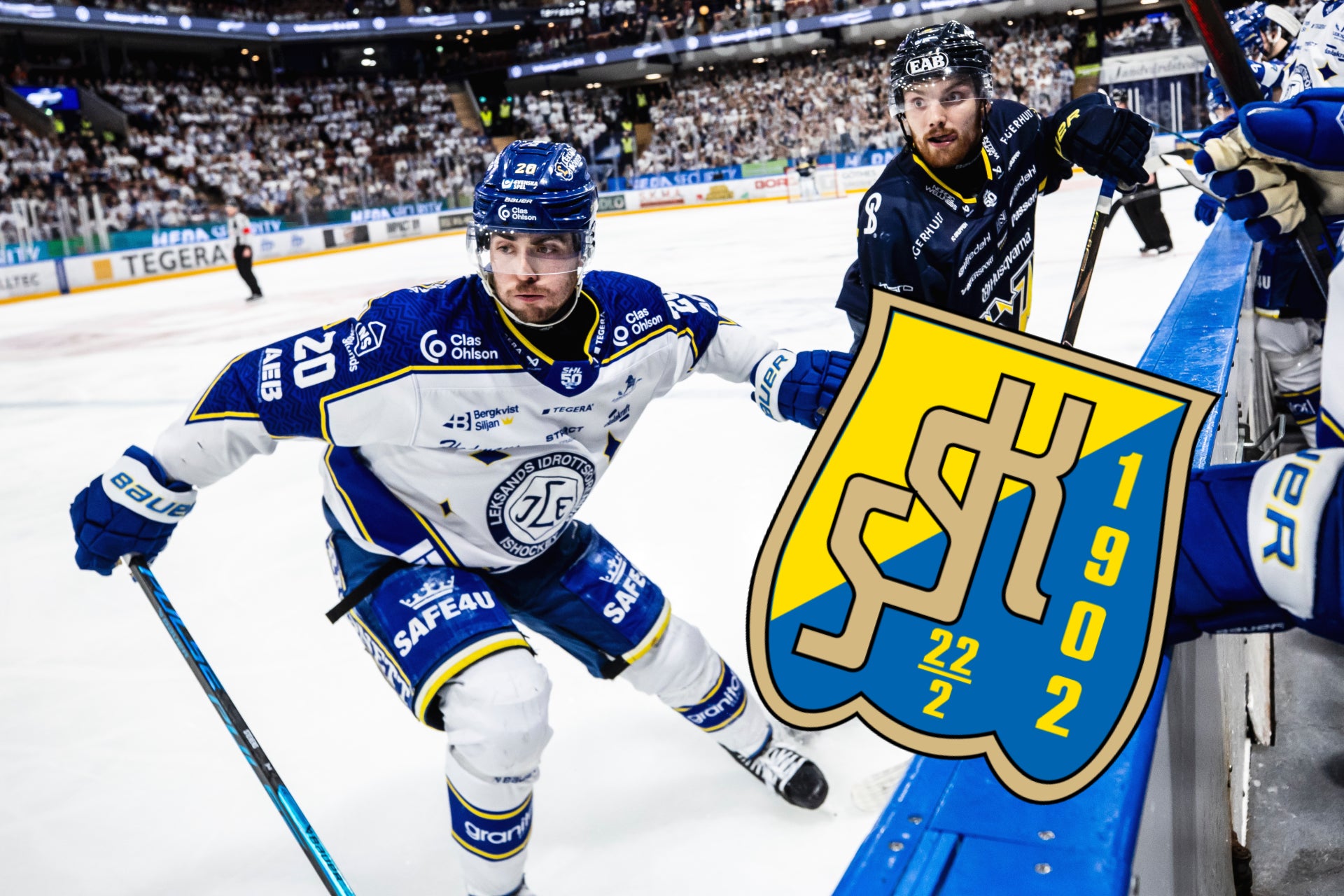 Ishockeymatch med spelare i blå och vit tröja som skär över isen nära banden med publikhall i bakgrunden.